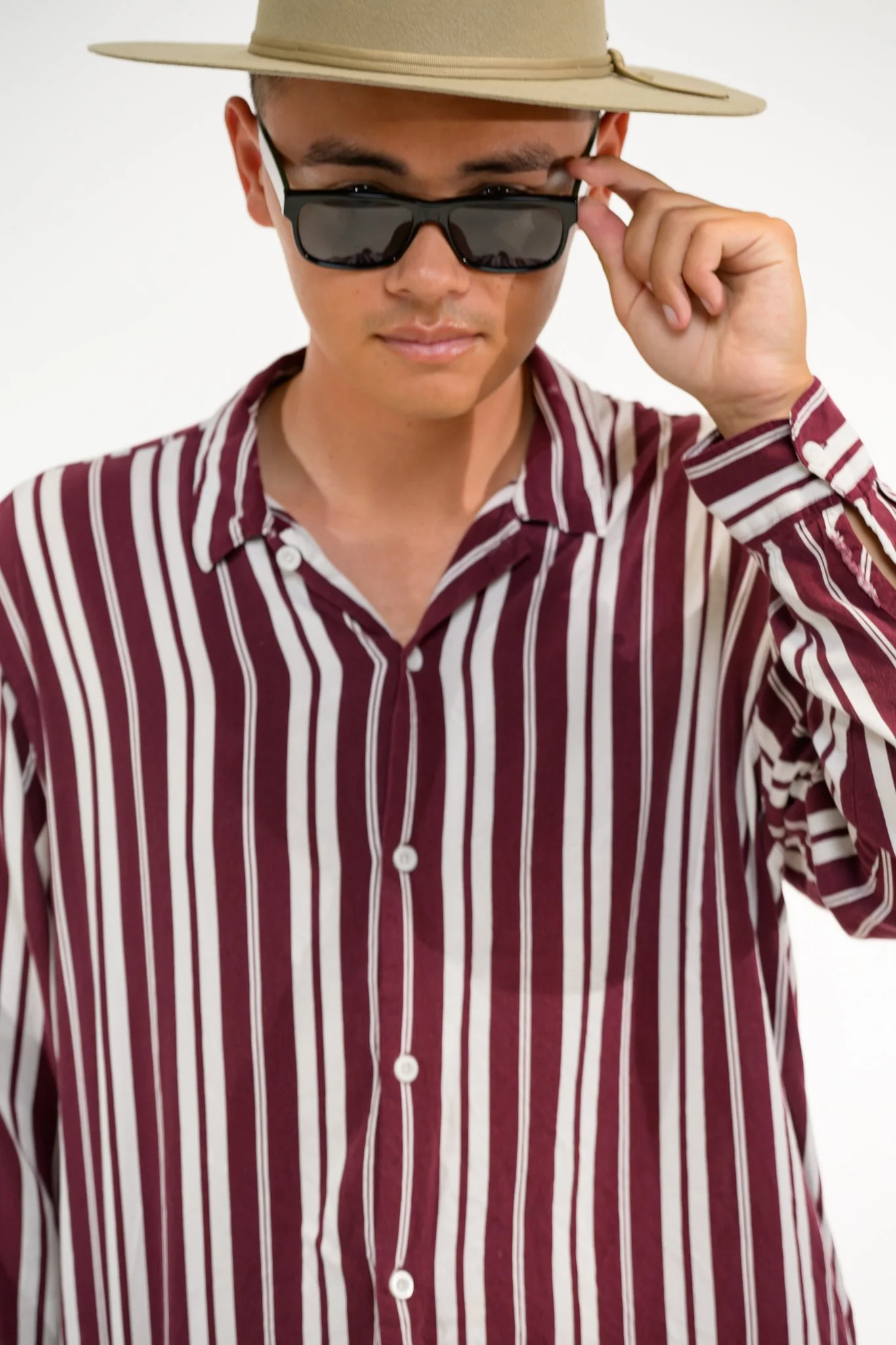 A man wearing a beige wide-brimmed hat, black sunglasses, and a maroon pinstriped button-up shirt.