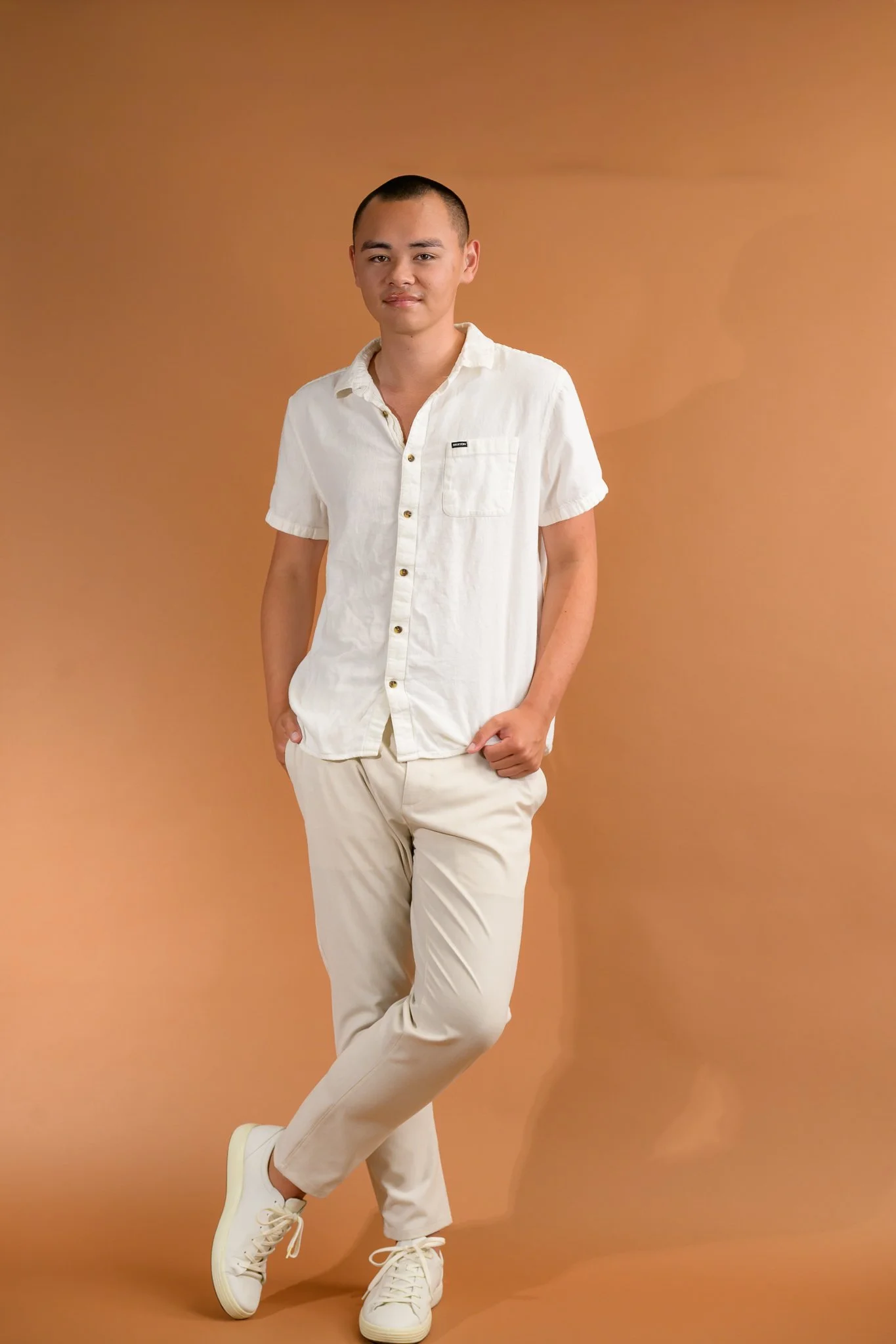 A young man with short hair dressed in a white short-sleeve shirt and beige pants, standing against a plain brown background with one leg crossed in front of the other and hands casually resting in his pockets.
