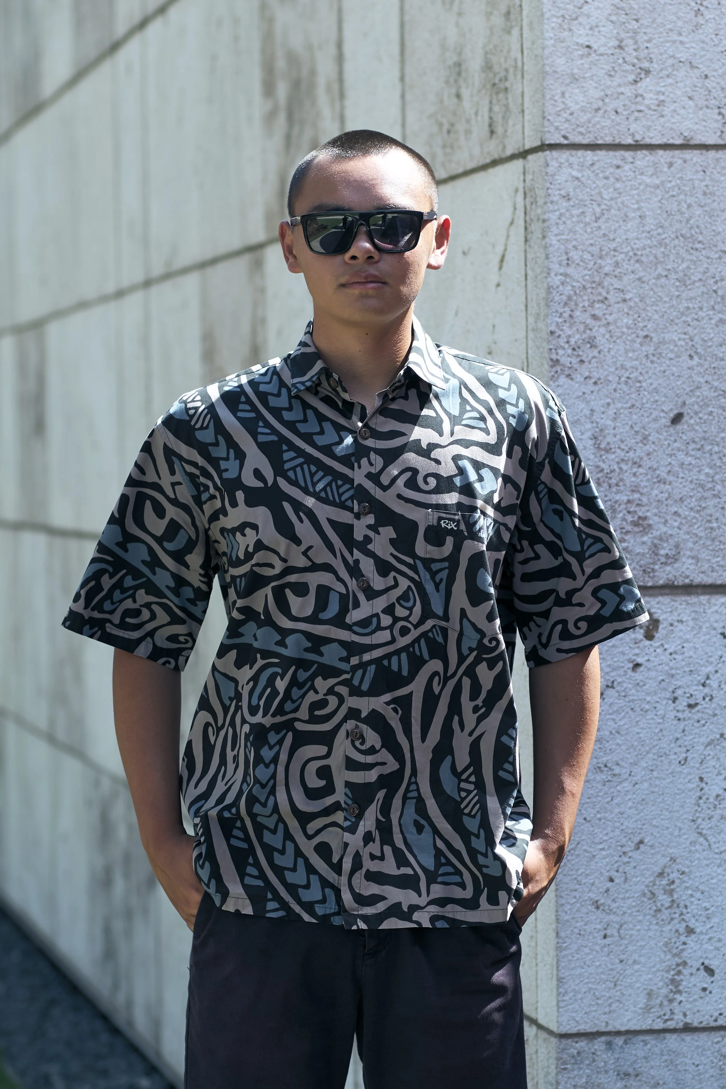 Young man with short hair and sunglasses, wearing a patterned short-sleeve shirt, standing outdoors against a textured stone wall.