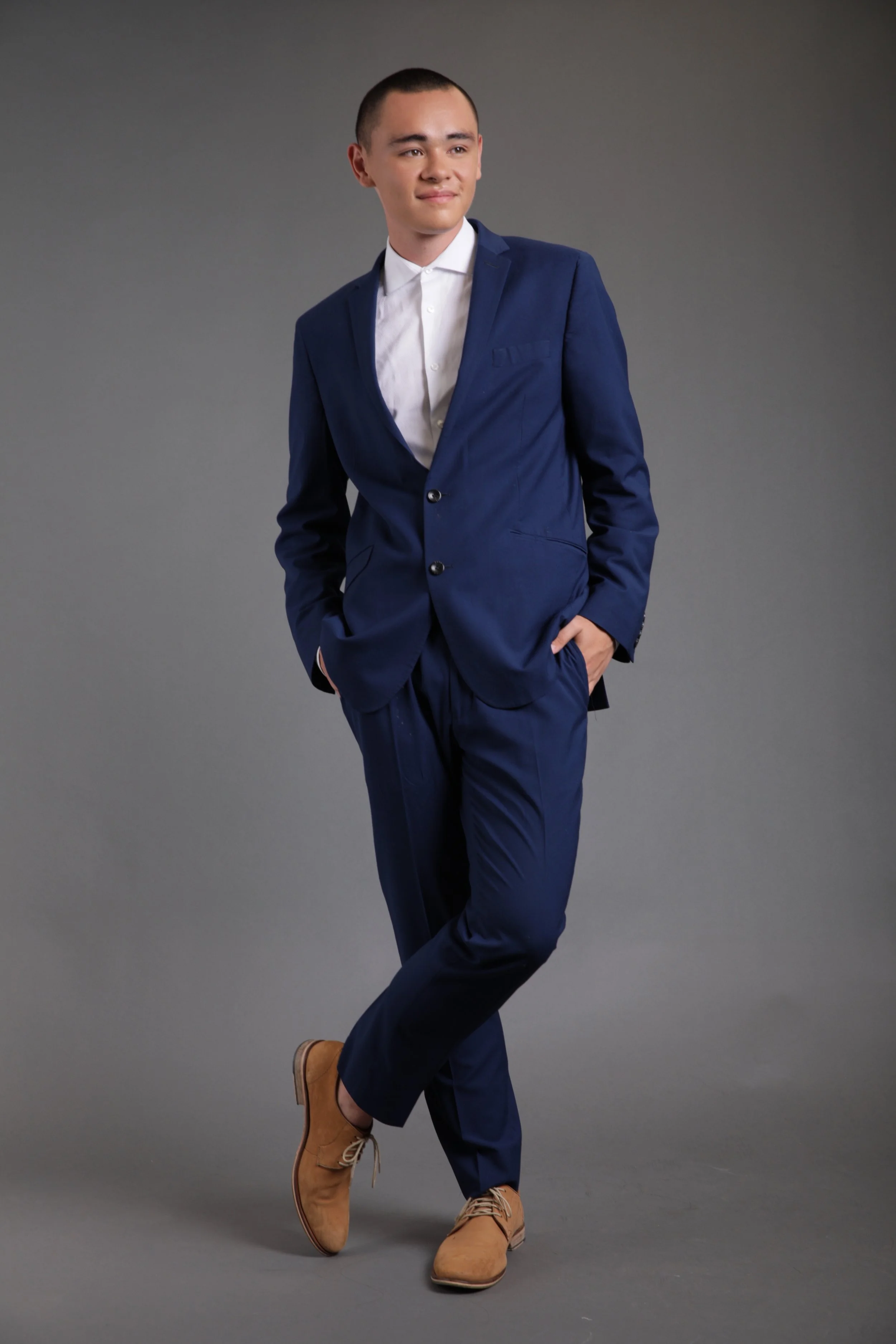 A young man with short hair wearing a navy blue suit and white shirt, tan boots, posing with hands in pockets against a gray background.