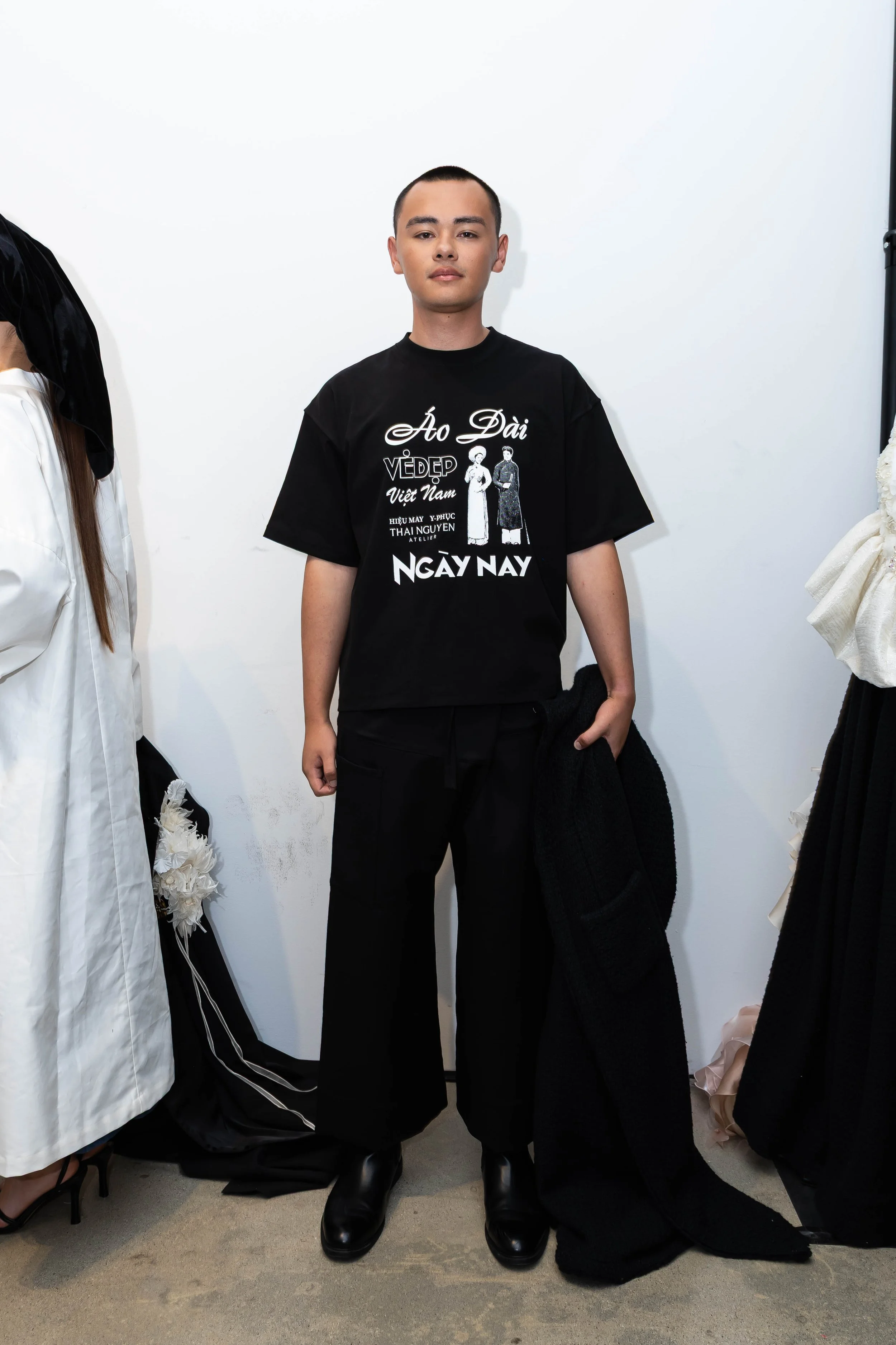 Young man with short hair standing against a white wall, wearing a black T-shirt with Vietnamese text and illustrations, black pants, and black shoes, holding a black jacket in his left hand.