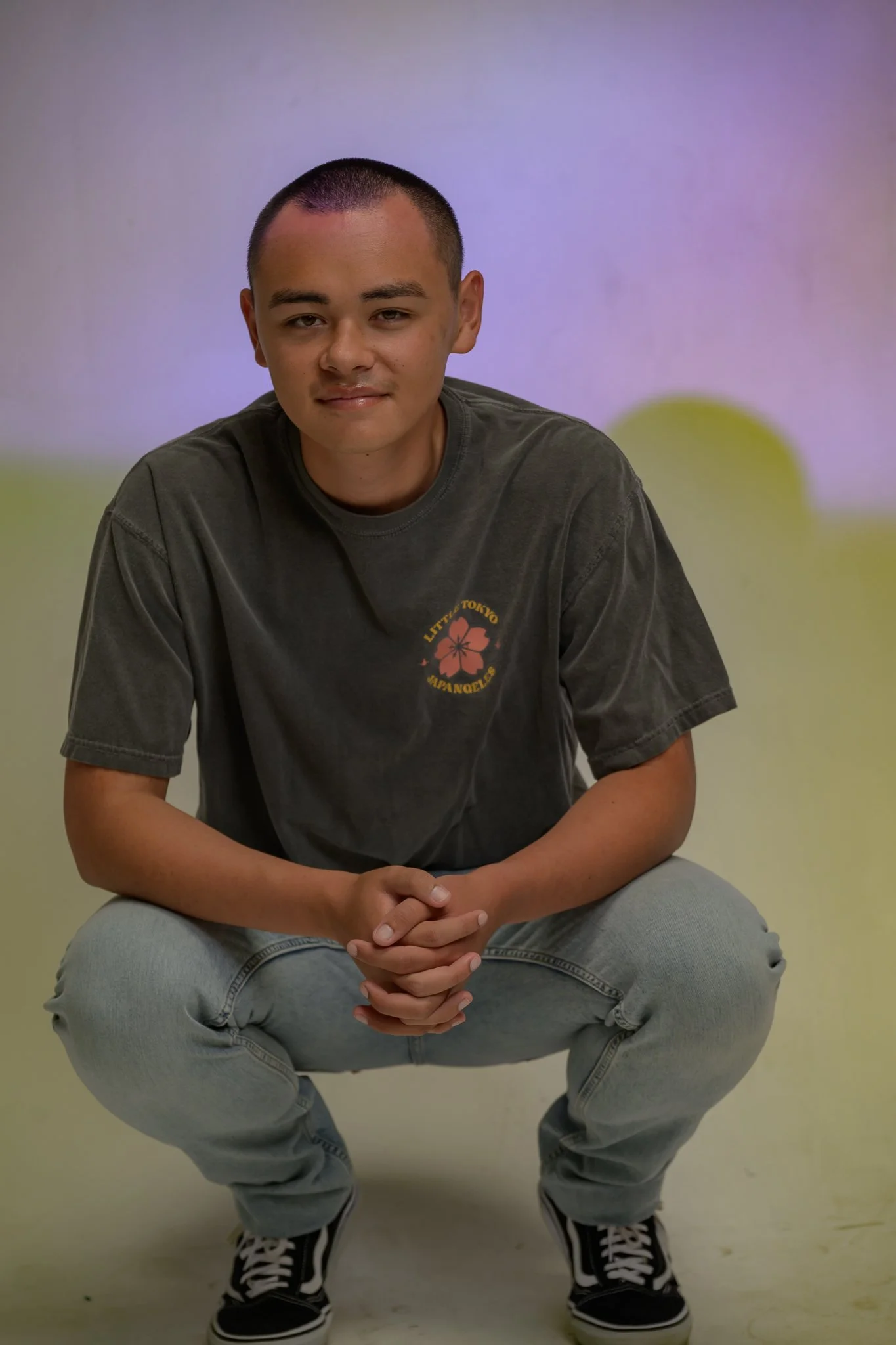 A young man with short hair and a slight smile, wearing a dark gray t-shirt with a graphic, light gray jeans, and black sneakers with white laces, squatting on a light-colored floor against a plain multicolored background.