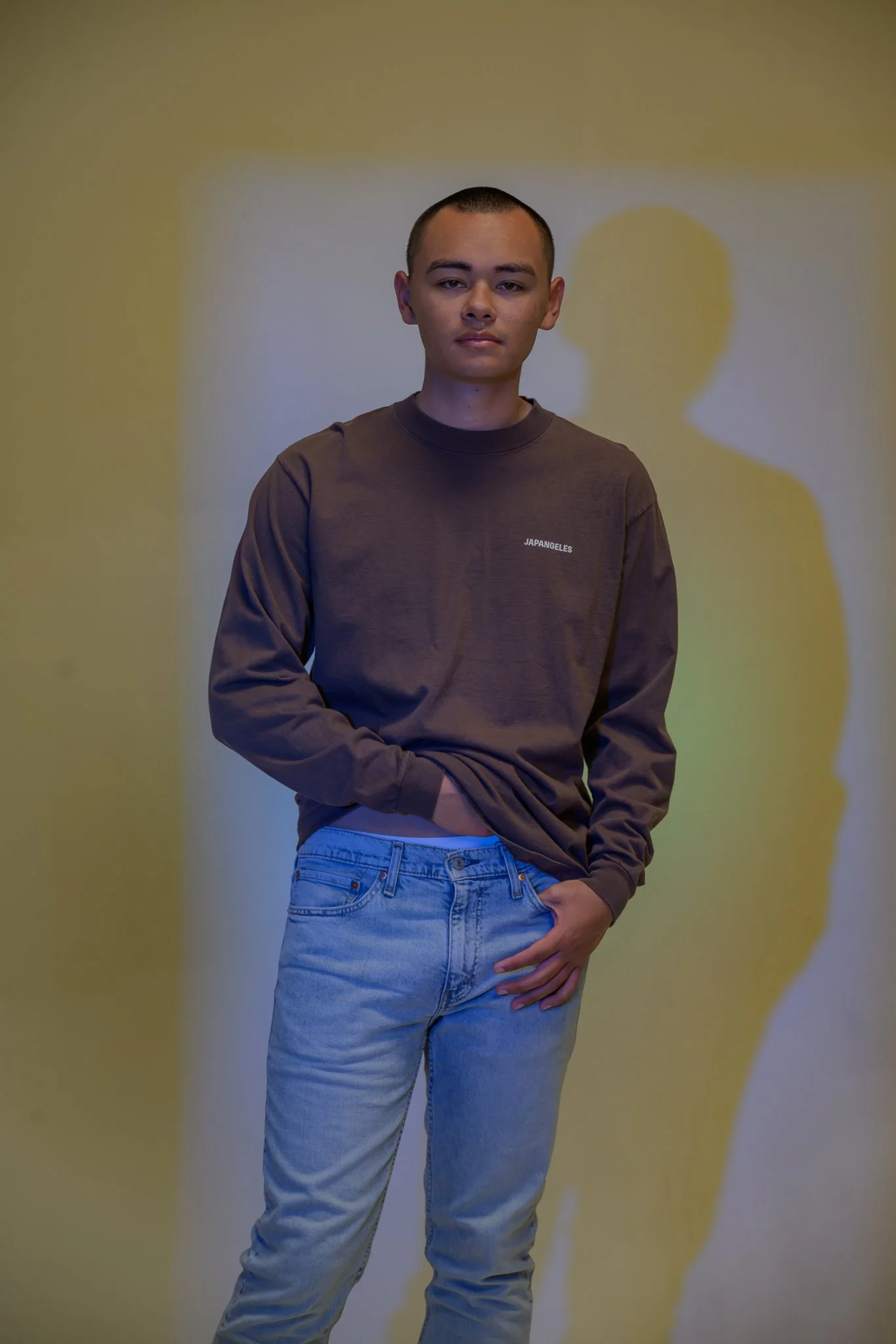 Young man with short dark hair, wearing a brown sweatshirt with white text that says 'JAPANGEL' and light blue jeans, standing in front of a yellow wall with a shadow of his profile.