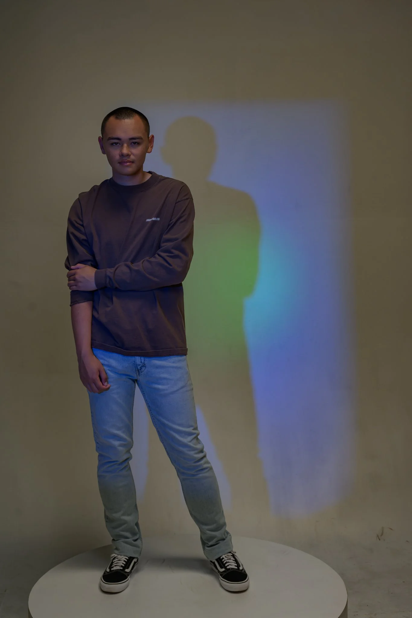 A young man with short hair stands with his arms crossed in front of a plain beige wall. He wears a gray sweatshirt, light blue jeans, and black-and-white sneakers. A shadow of his head is cast on the wall behind him.