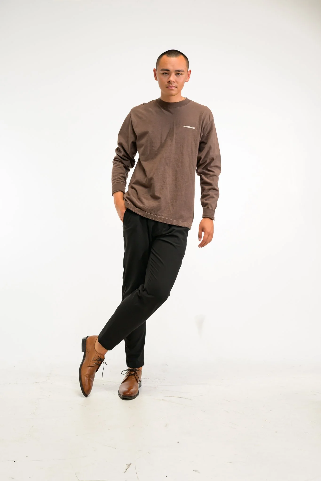 A young man with short hair wearing a brown long-sleeve shirt, black pants, and brown shoes standing against a plain white background.