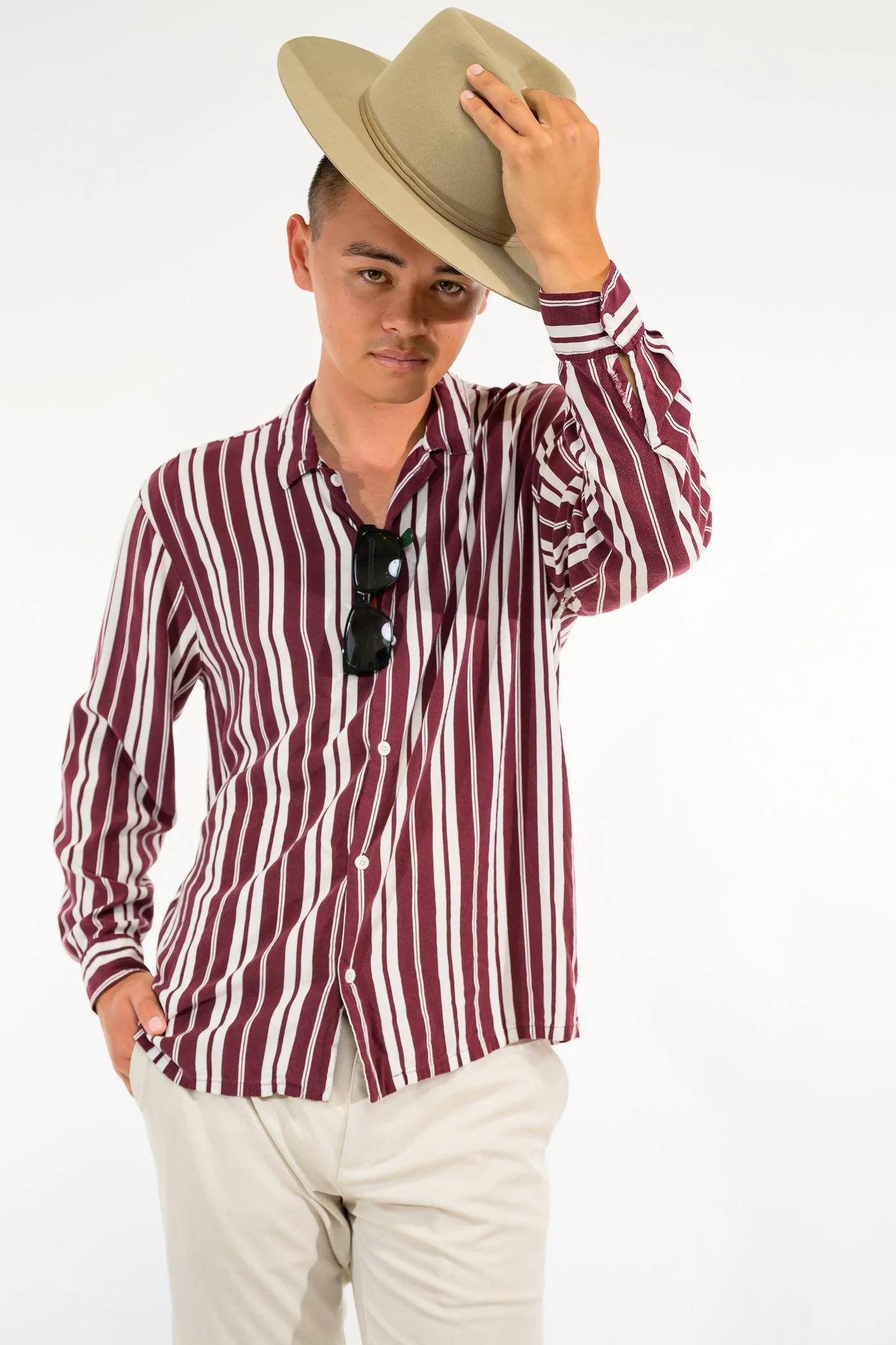 A young man wearing a beige hat, a red and white striped shirt, and beige pants, posing with one hand on his hip and the other adjusting his hat, against a plain white background.