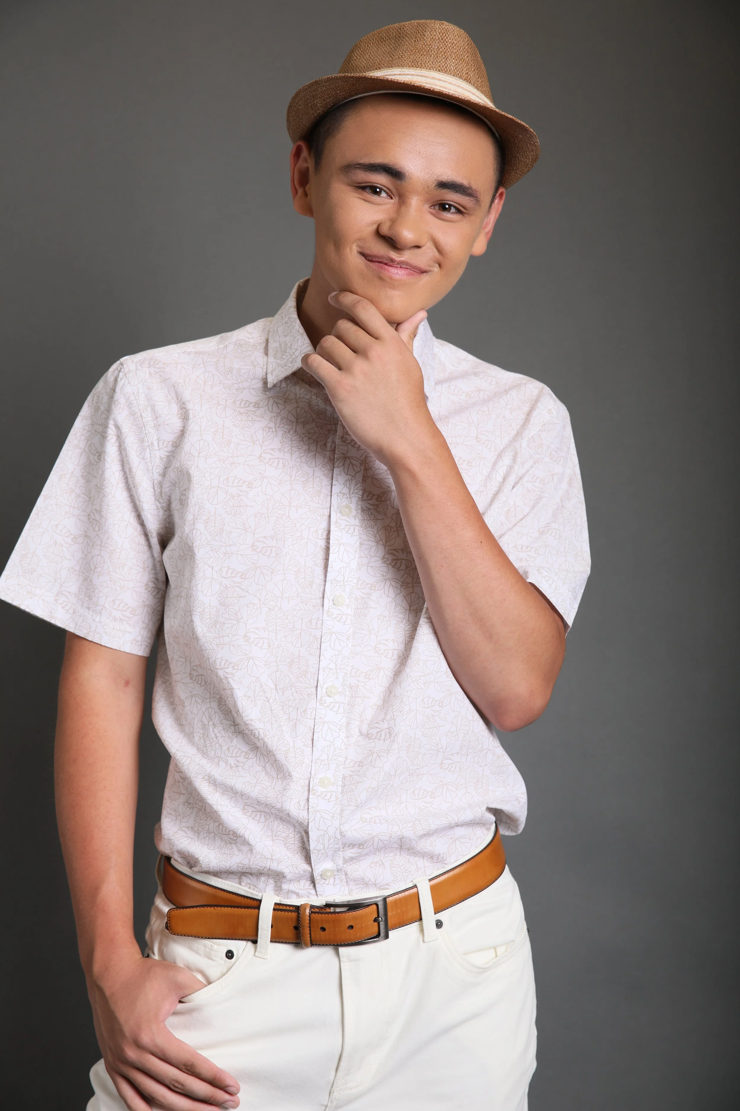 A young man dressed in a short-sleeved, light-colored patterned shirt, white pants, and a brown belt, wearing a tan hat, standing against a gray background, smiling slightly with his right hand resting on his chin and his left hand in his pocket.