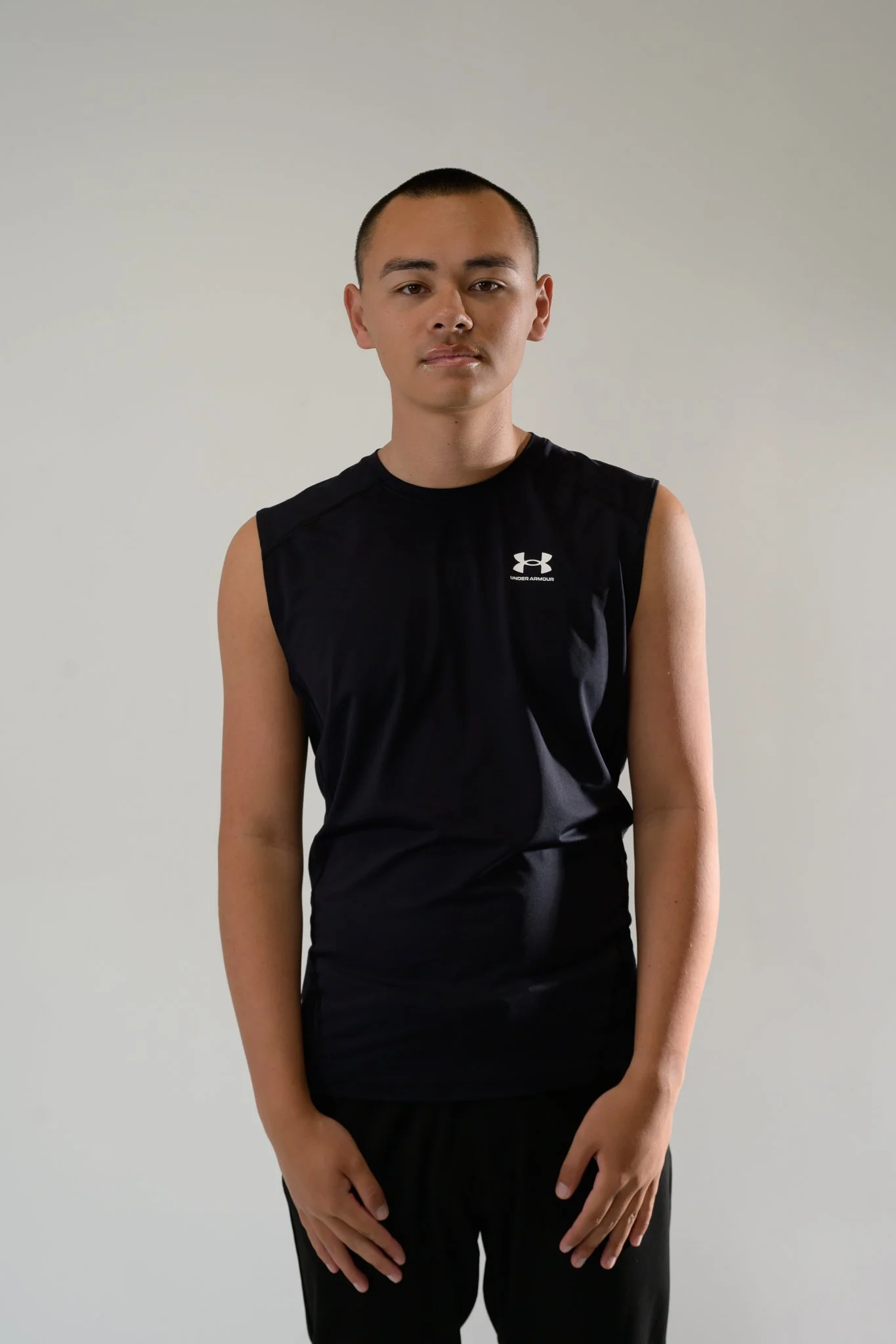 A person with a buzz cut hairstyle, wearing a sleeveless black Under Armour shirt and black pants, standing against a plain light gray background.