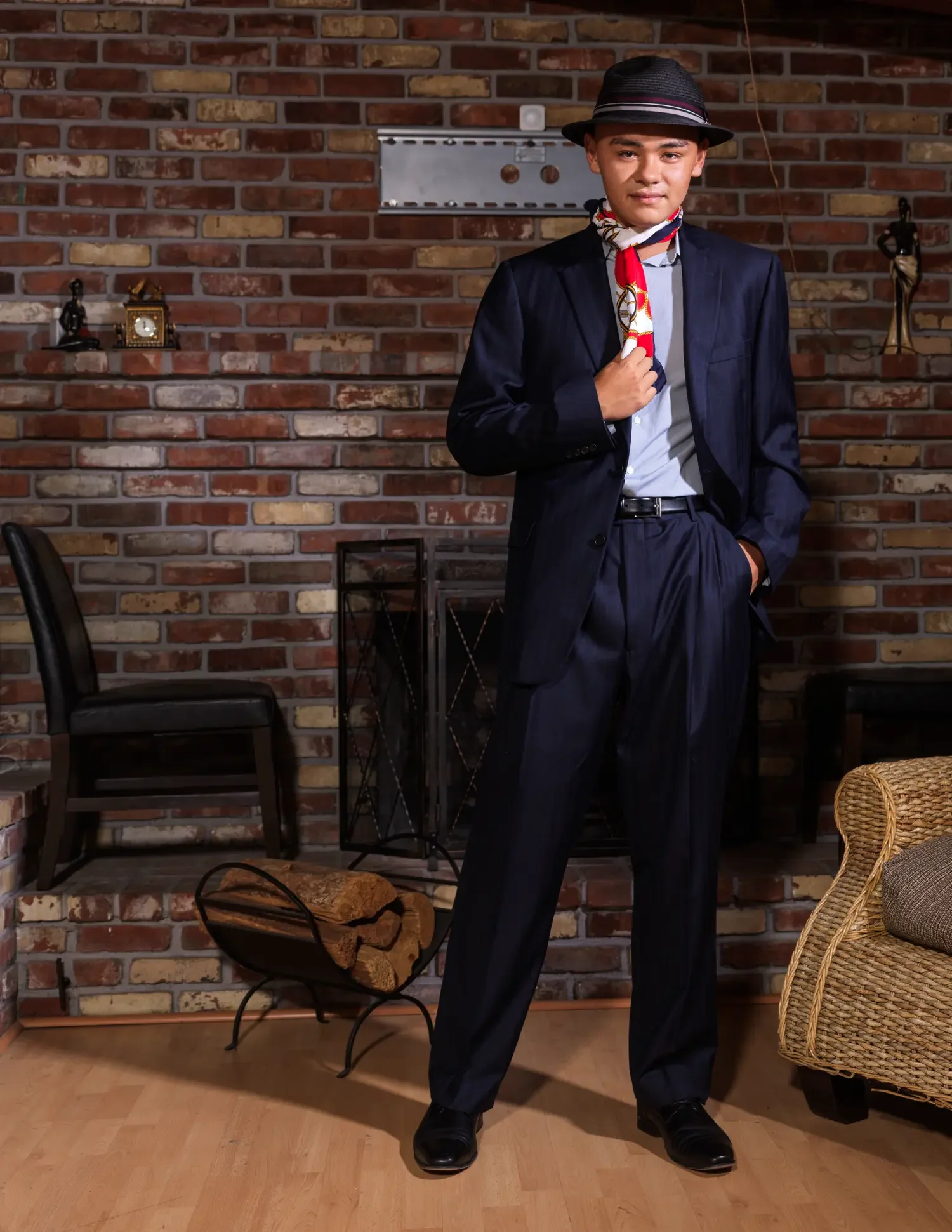 A young man dressed in a dark suit, light blue shirt, wearing a black fedora hat with a red, white, and blue striped band, and a patterned scarf around his neck, standing in a room with a brick wall. The room has a fireplace with logs, a black chair, and decorative statues and objects on the mantel.