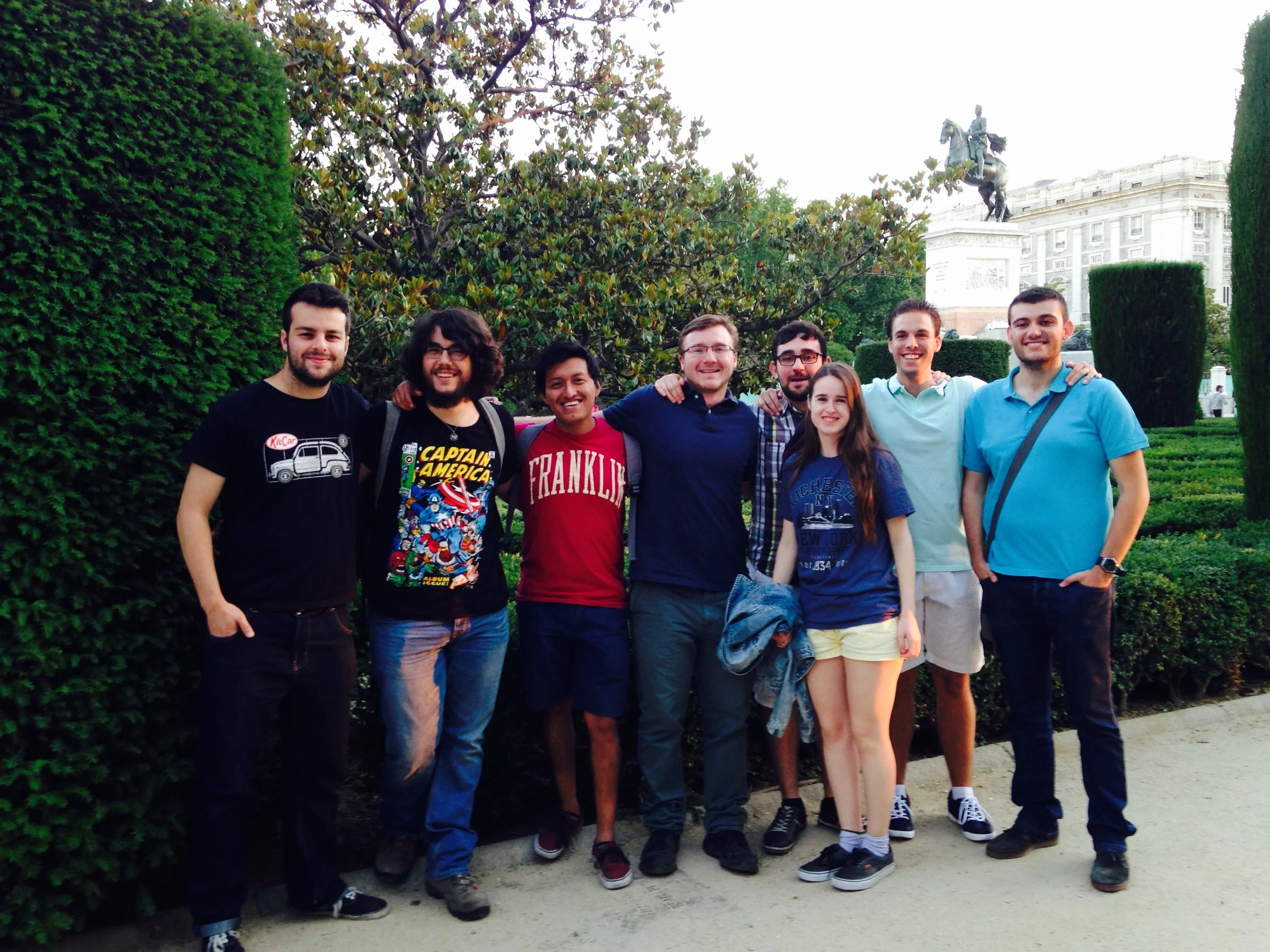 A group of nine young adults standing together outdoors, smiling for a photo. They are surrounded by green bushes and trees, with a historic building and a statue of a person on horseback seen in the background.