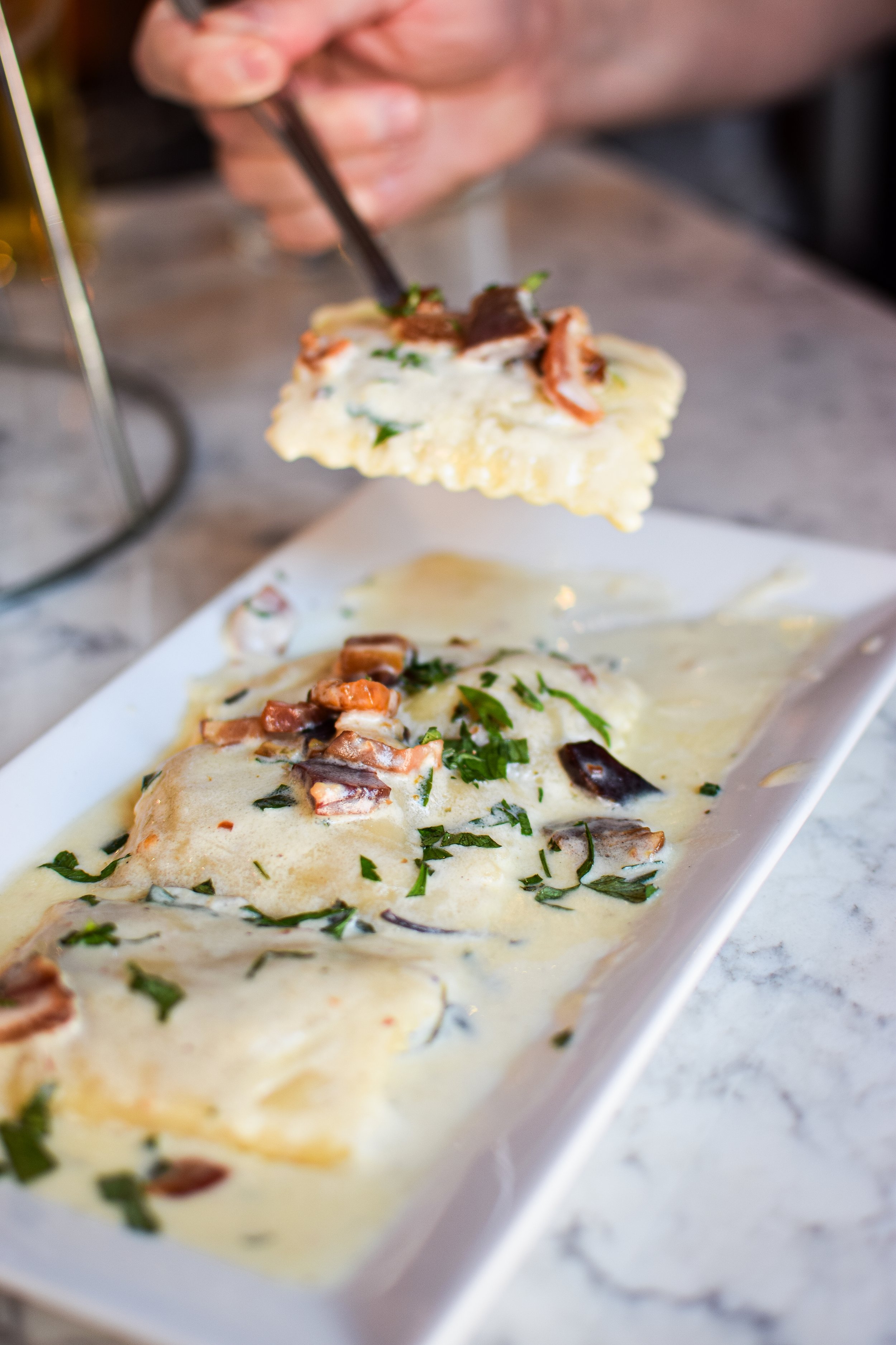 Close-up of a hand using tongs to serve pumpkin-stuffed ravioli topped with cream sauce, herbs, and pancetta from a white rectangular platter on a marble surface.