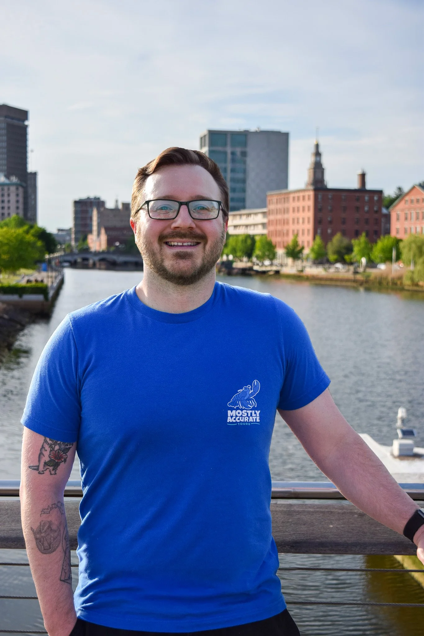 A smiling man with glasses and tattoos on his arms, wearing a blue t-shirt with a logo, stands on a bridge near a river, with city buildings and trees in the background.