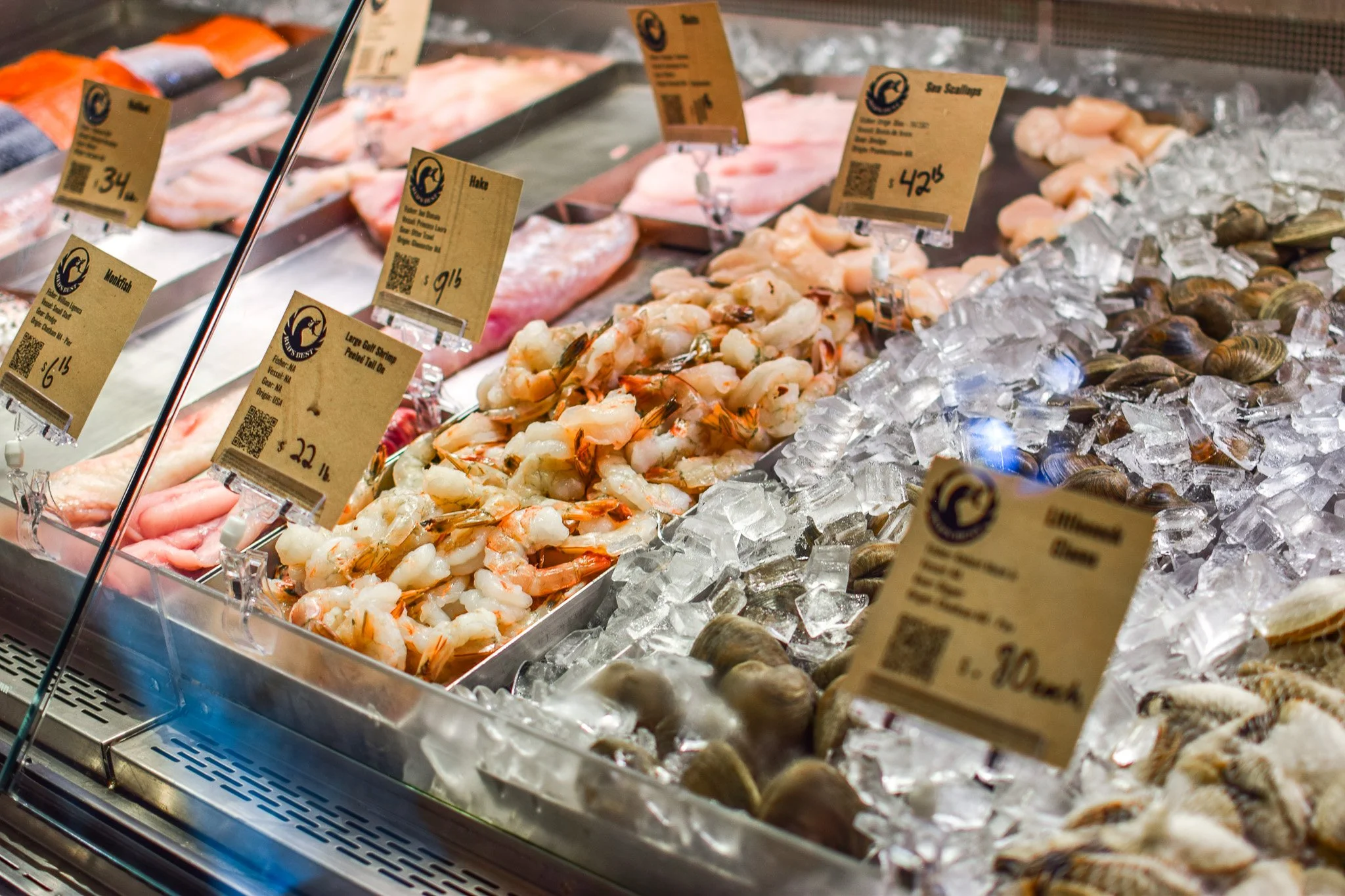 A display case with various fresh seafood including shrimp, scallops, fish fillets, and shellfish on crushed ice, with price tags and labels.