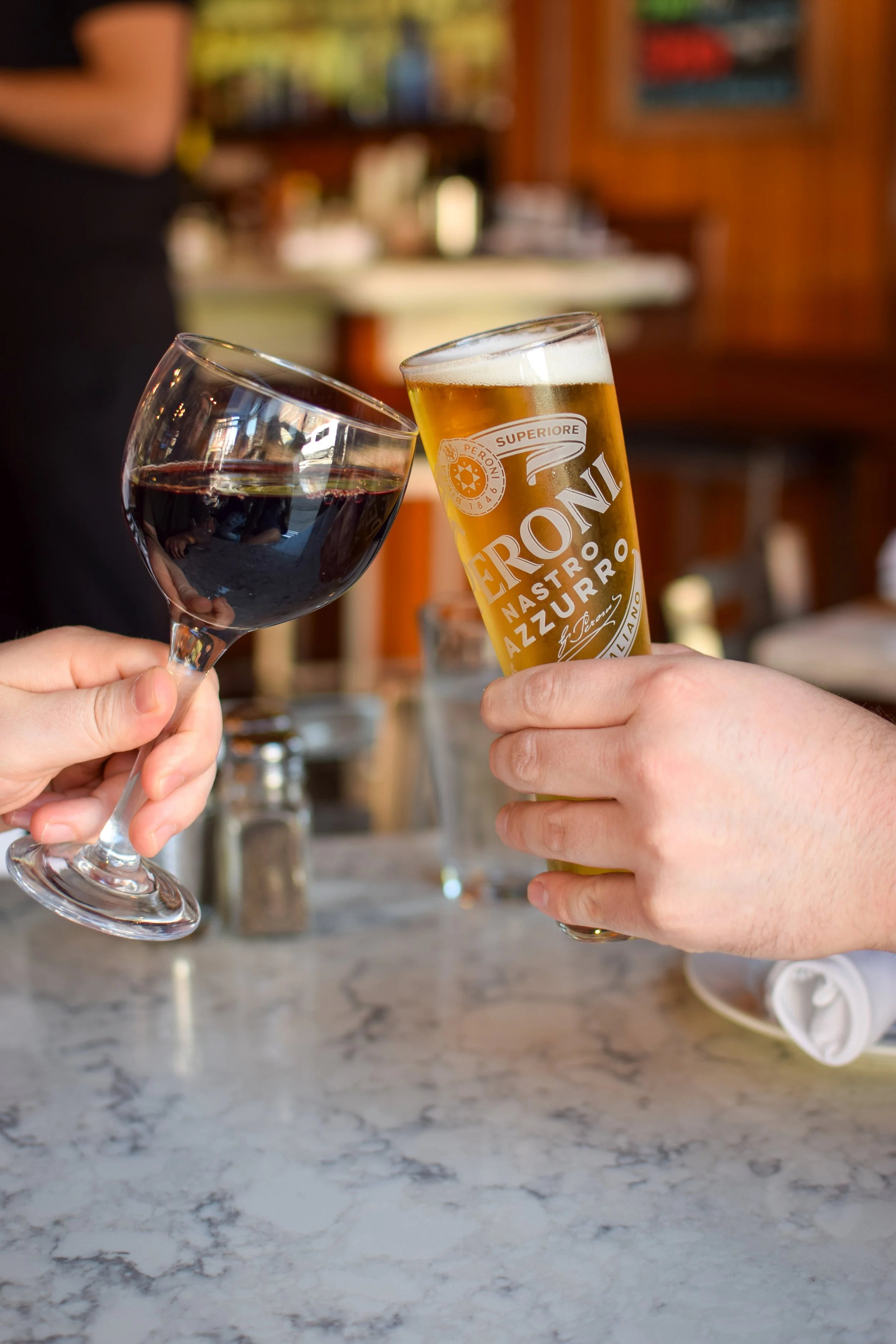 Two people holding wine glass and beer glass clinking together in a toast, with a bar or restaurant setting in the background.