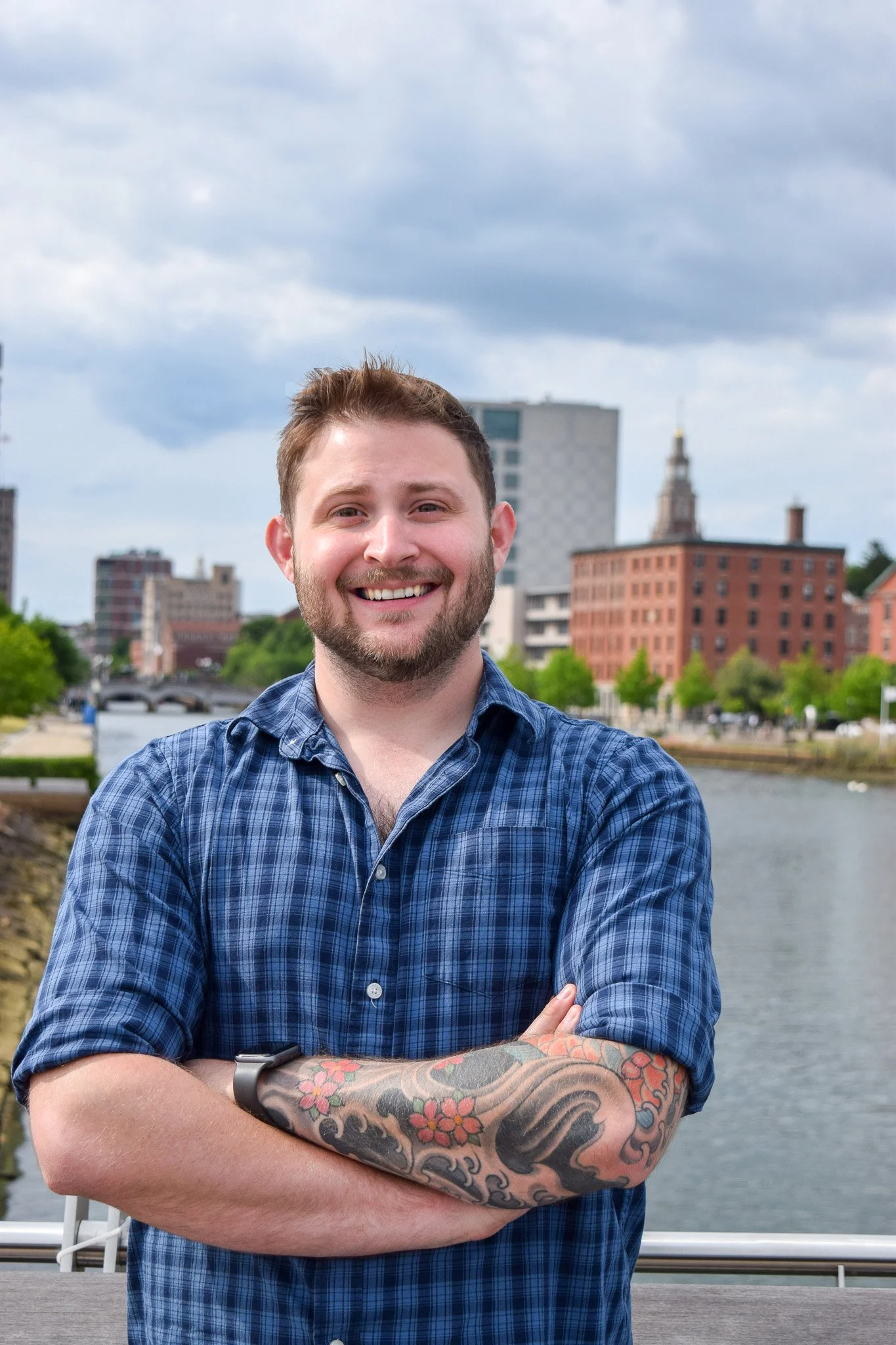 A smiling man with short hair and a beard standing outdoors near a body of water, with a city skyline and cloudy sky in the background, arms crossed, wearing a blue plaid shirt with tattoos on one arm.