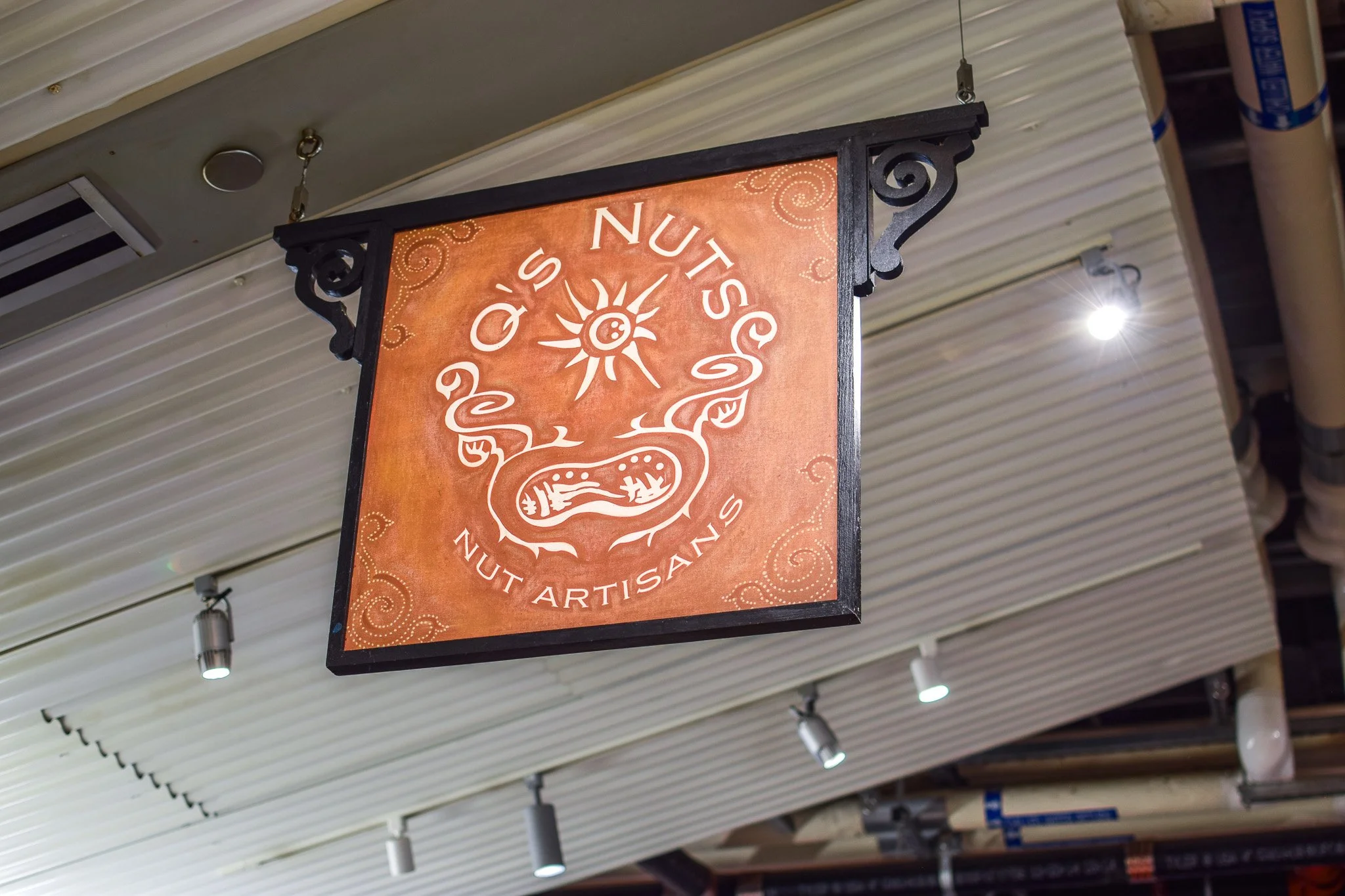 Hanging sign for Q's Nuts shop or product, featuring an artistic nut or acorn design on an orange background with decorative swirl patterns.
