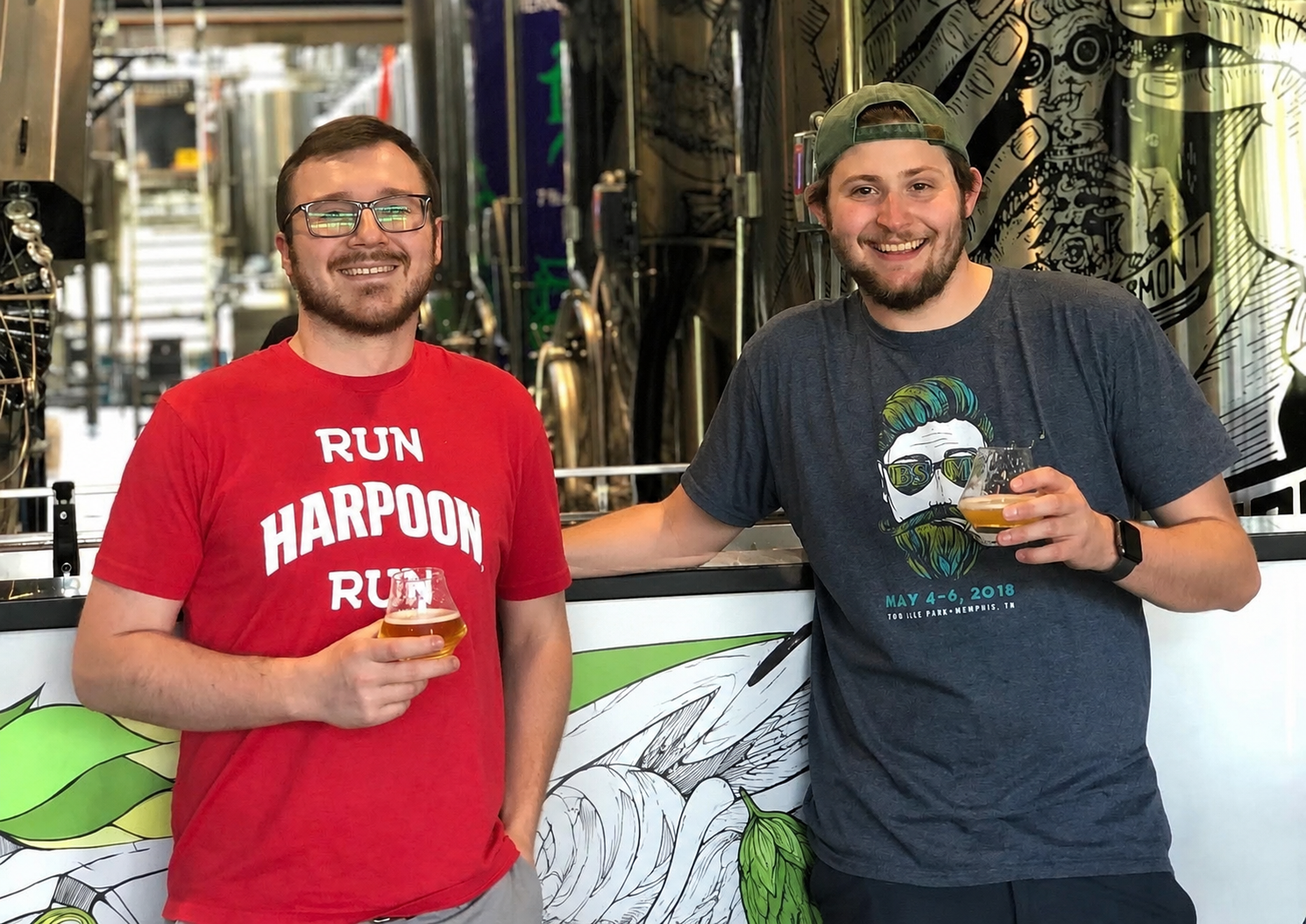 Two men smiling and holding glasses of beer at a brewery or bar. One man is wearing a red T-shirt with white text, and the other is wearing a dark gray T-shirt with a graphic and text. They are standing in front of a mural or artwork with brewing equipment visible in the background.