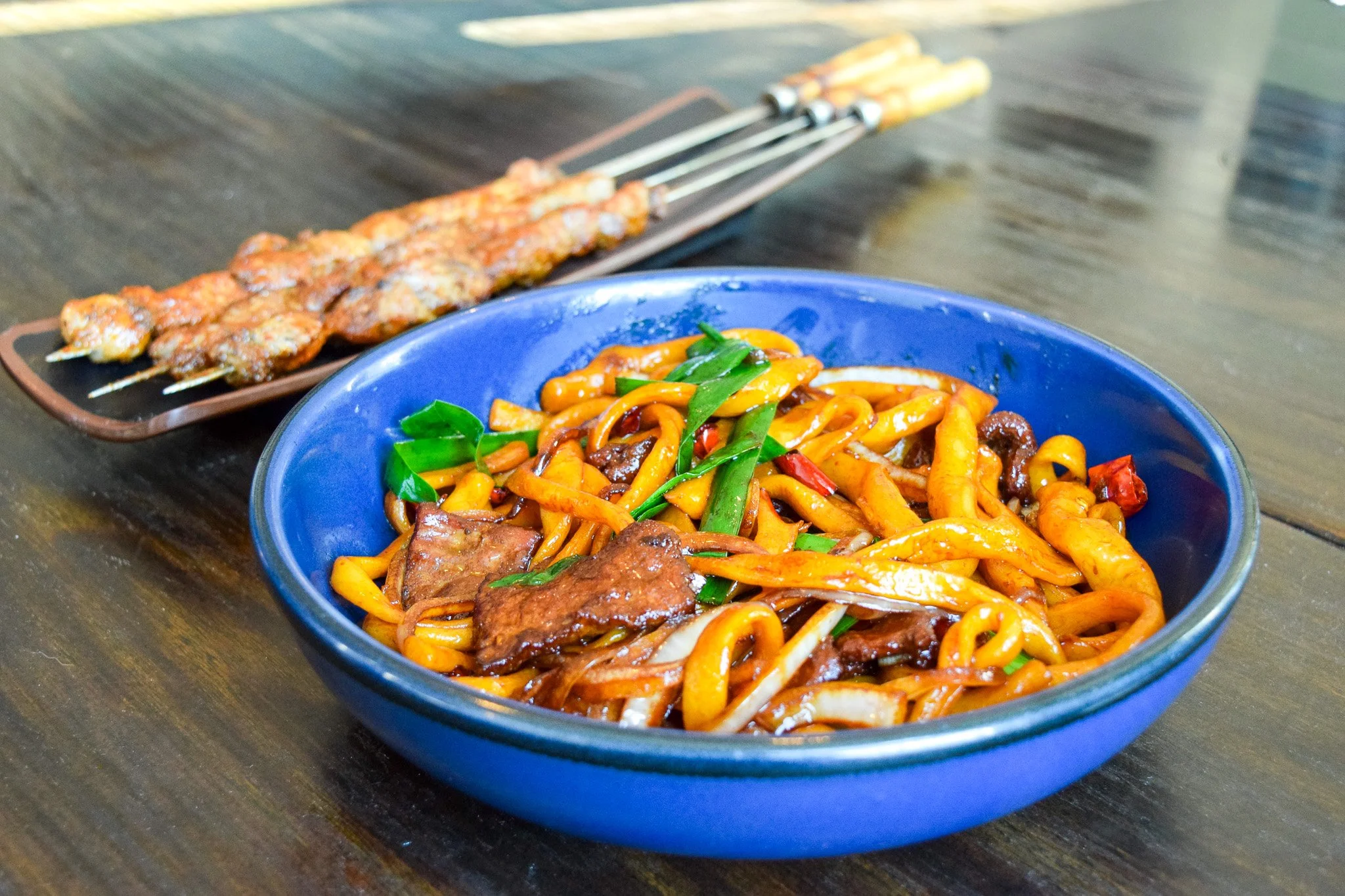 A blue bowl filled with stir-fried noodles with vegetables and pieces of meat. In the background, a plate with grilled skewers on a wooden table.