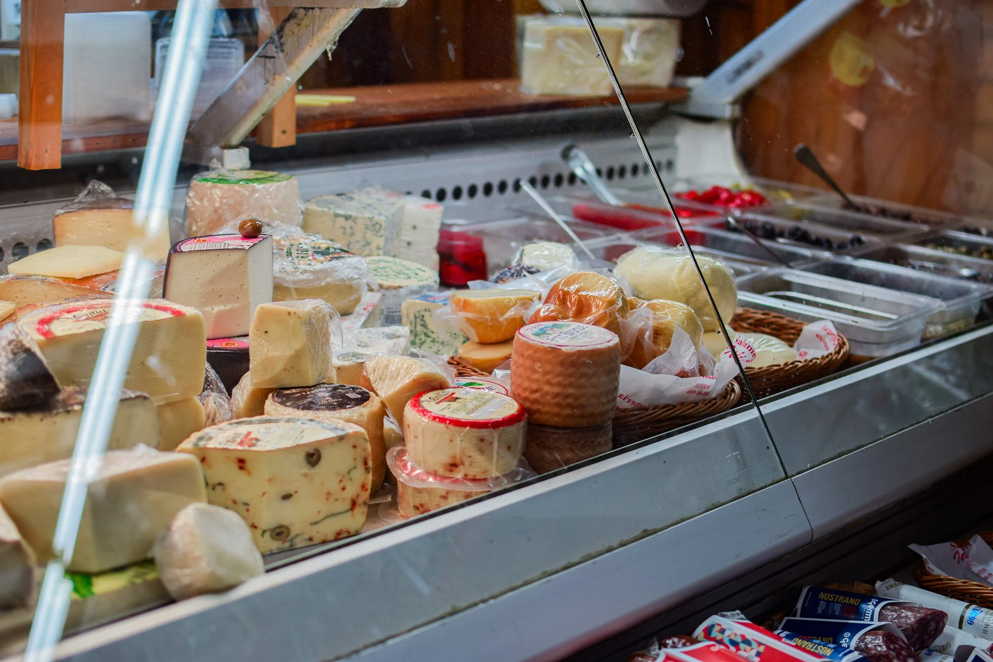 A display case filled with various types of cheeses, some wrapped in plastic, with a wooden shelf and other deli items in the background.