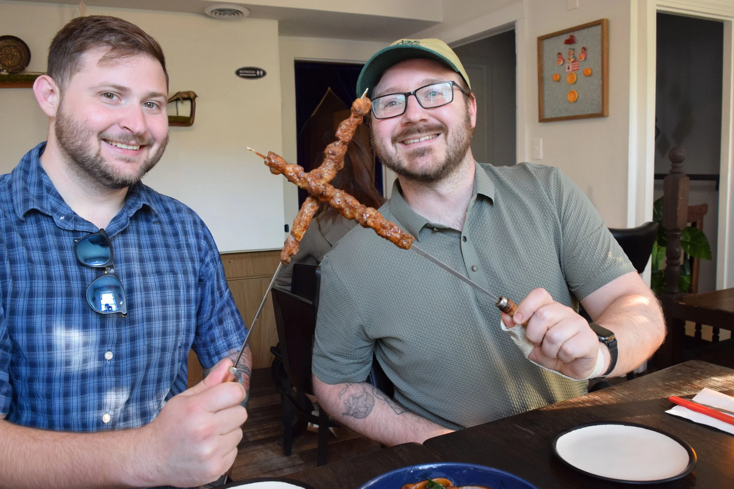 Two men smiling, sitting at a table, holding skewers of grilled meat, with a casual indoor setting in the background.