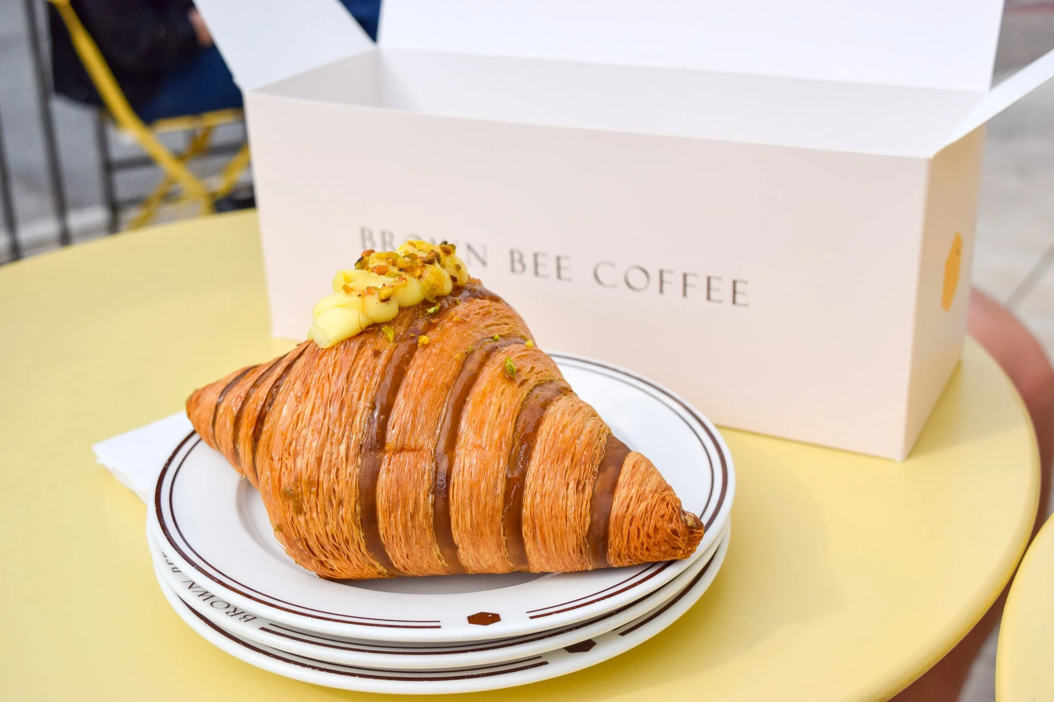 A croissant pastry topped with cream and crushed pistachios, placed on a white plate with a logo, in front of a white box with the words "BROWN BEE COFFEE," on a yellow table.