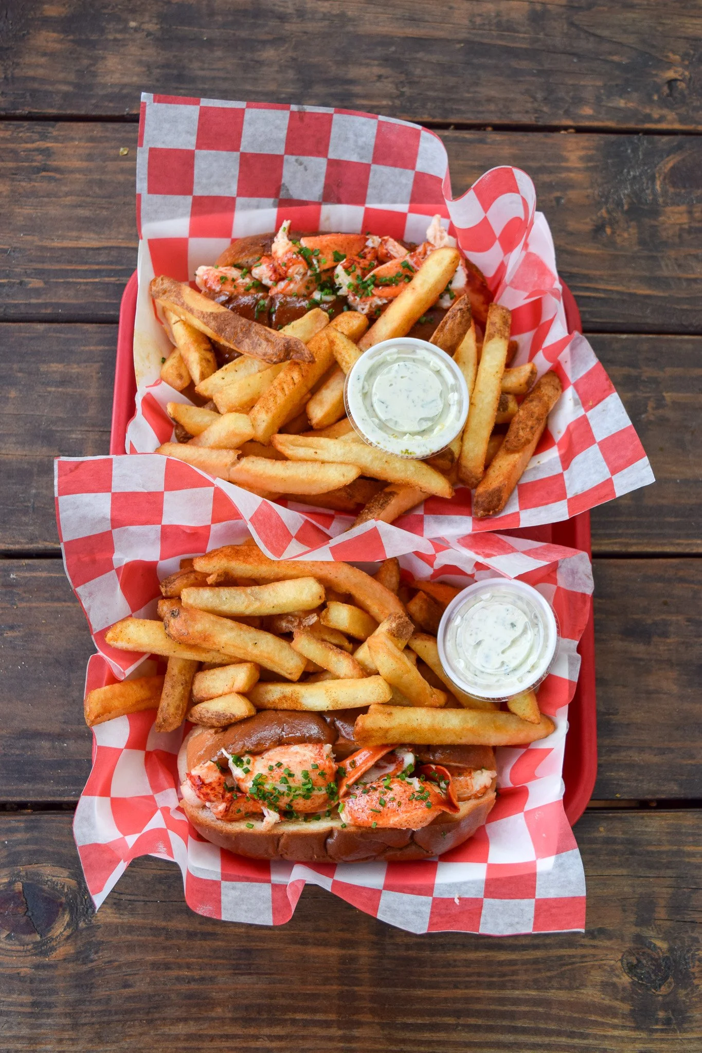 Two baskets of Dune Brother's Lobster Rolls and French fries on a rustic wooden table, with each basket lined with red and white checkered paper and a small container of dipping sauce.