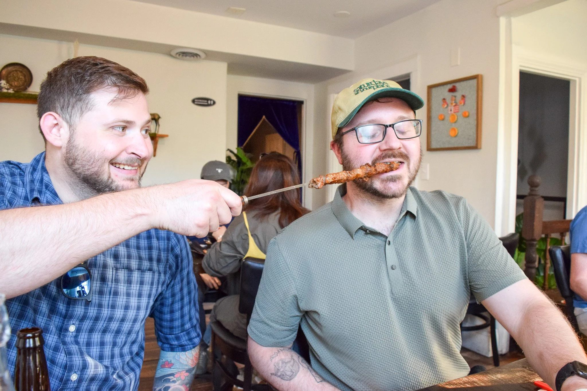 Two men at a social gathering. One man has short brown hair, beard, and is wearing a blue plaid shirt and sunglasses. The other man has glasses, a beard, a cap, and is wearing a gray-green shirt. The man in plaid is handing a skewer with meat to the man in glasses, who is about to eat it. The background shows a few other people, some furniture, and wall decorations in a home setting.