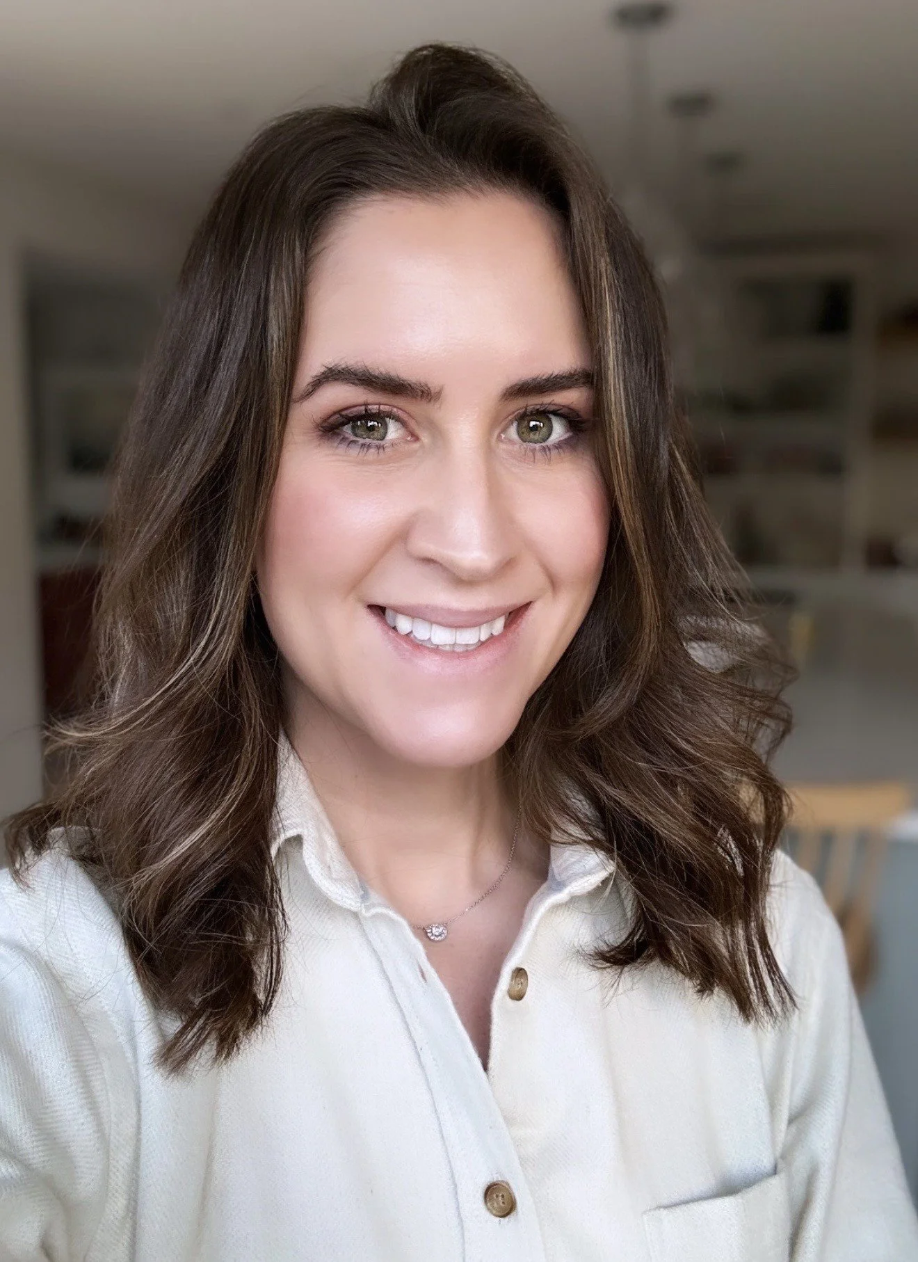 Close-up of a smiling woman with wavy brown hair, wearing a cream-colored button-up shirt and a necklace, in an indoor setting.