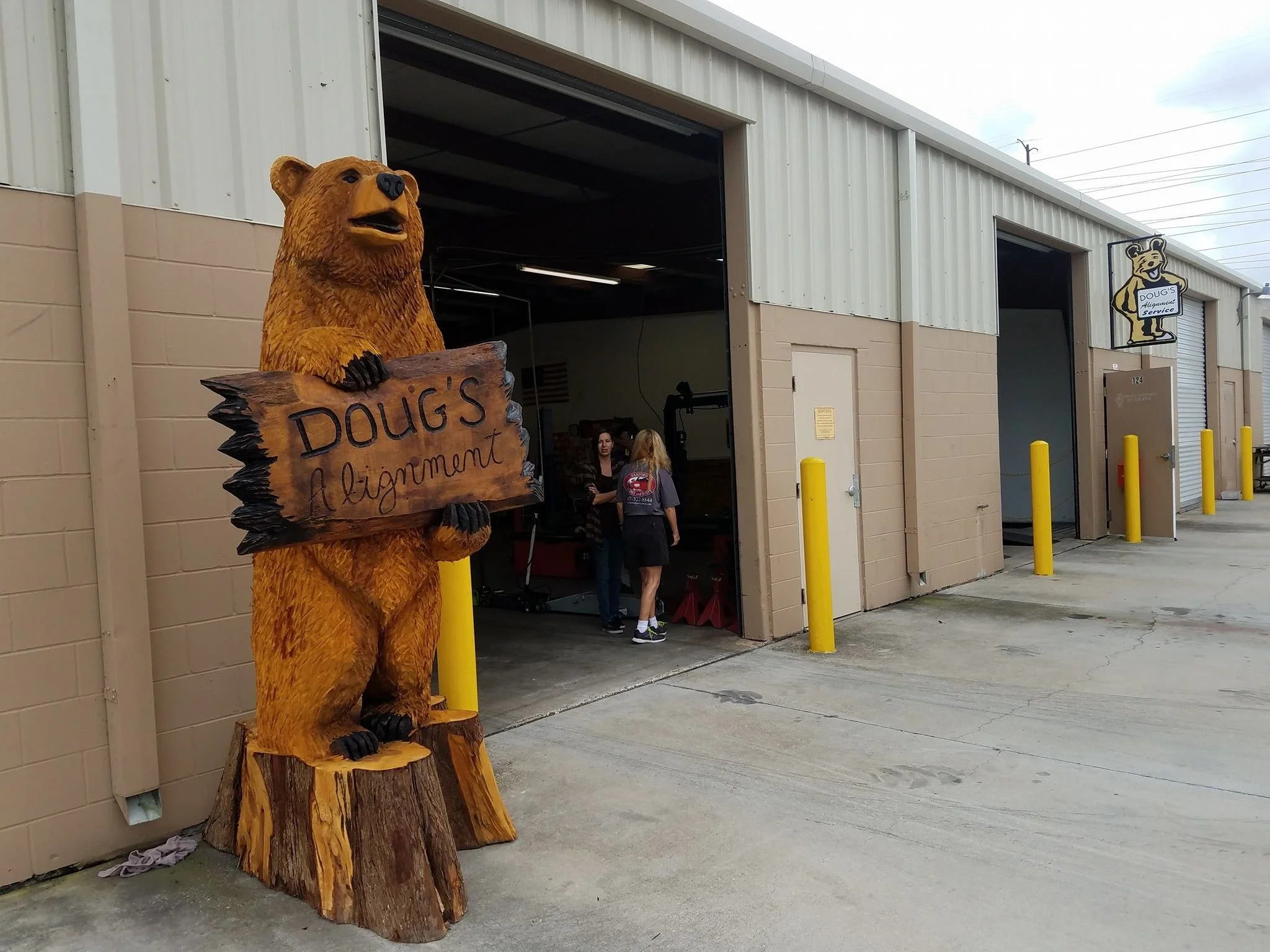 A wooden bear statue holding a sign that reads 'Doug's Alignment' outside a beige industrial building with open garage doors. Two women are standing inside the building near the entrance.