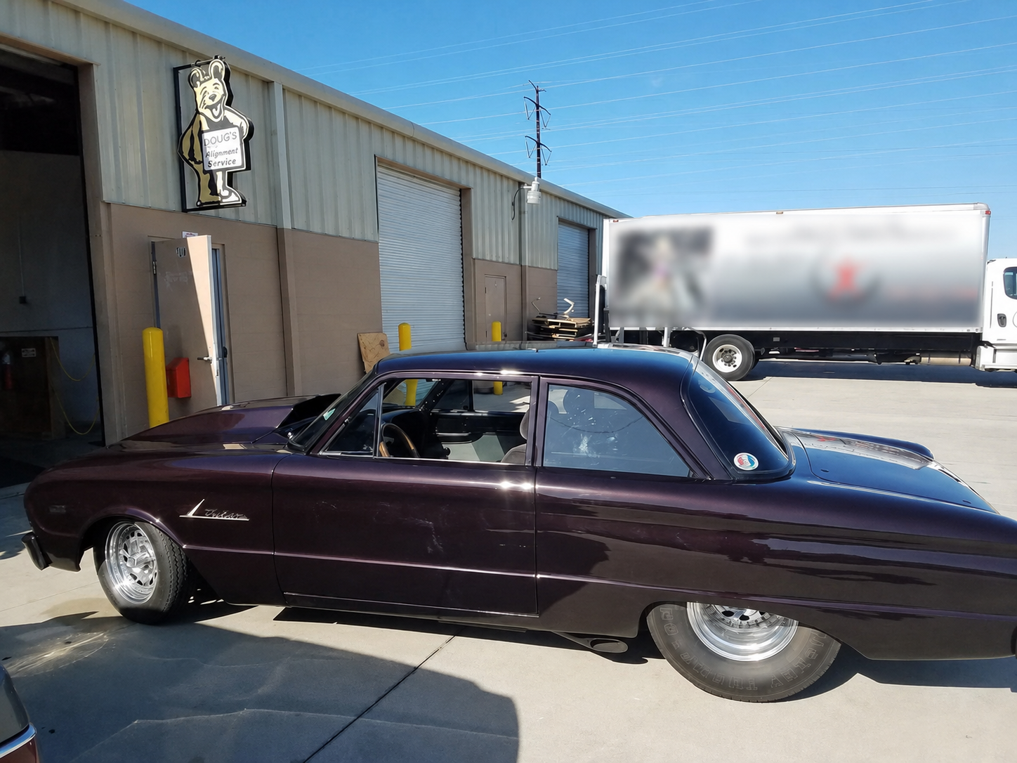 A vintage purple Chevrolet Corvette parked outside an auto repair building named Doug's Alignment Service. The building has a sign with a cartoon bear wearing a mechanic uniform. A large semi-truck with a blurred trailer is parked in the background under a clear blue sky.
