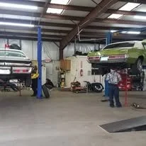 Automotive repair shop with two cars on lifts, mechanics working underneath, and tools scattered around.