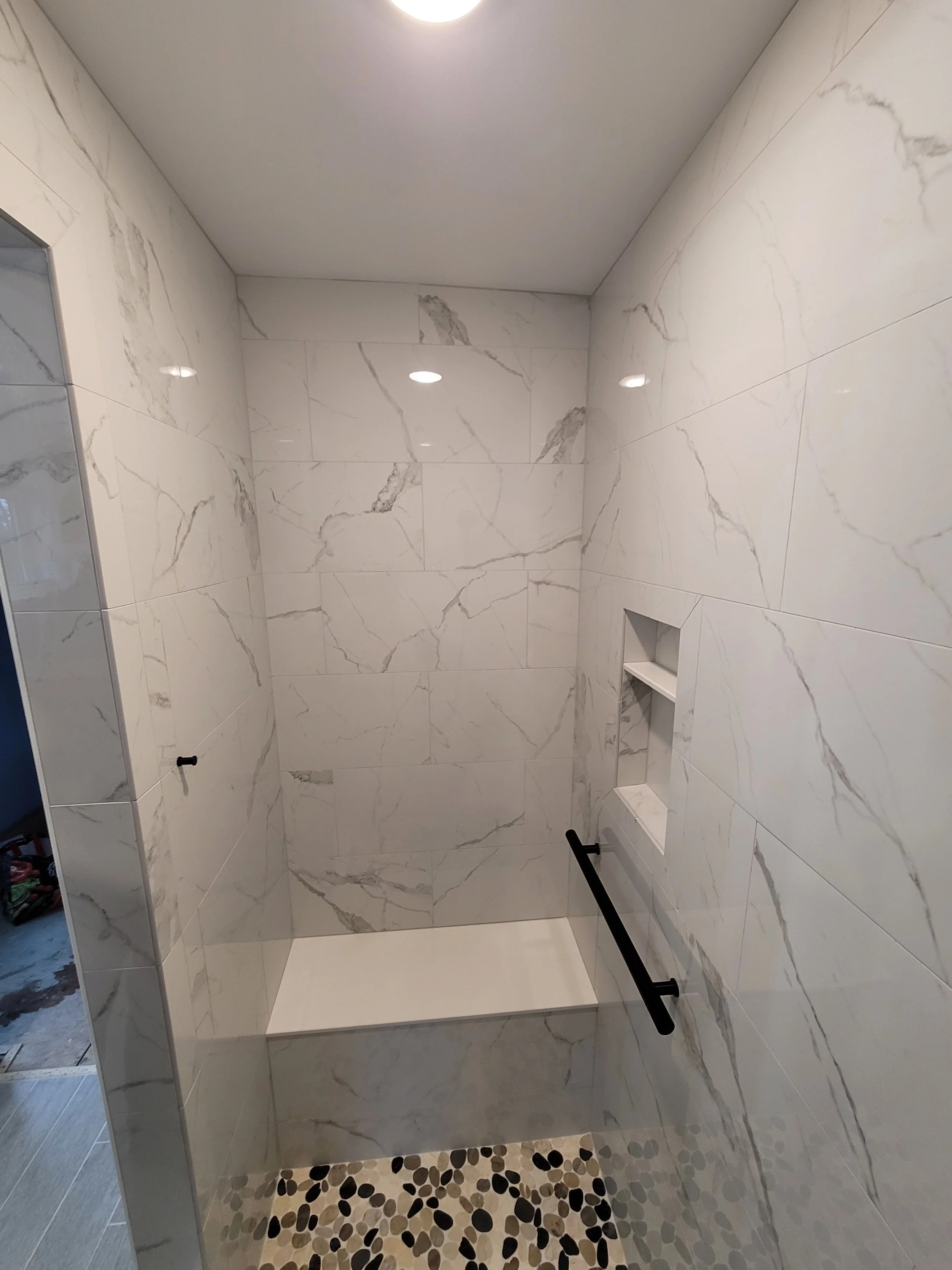A shower stall with white marble tiles on the walls and a pebble-patterned tile on the floor. There is a built-in shelf in the wall and black horizontal grab bars. The ceiling has a light fixture.