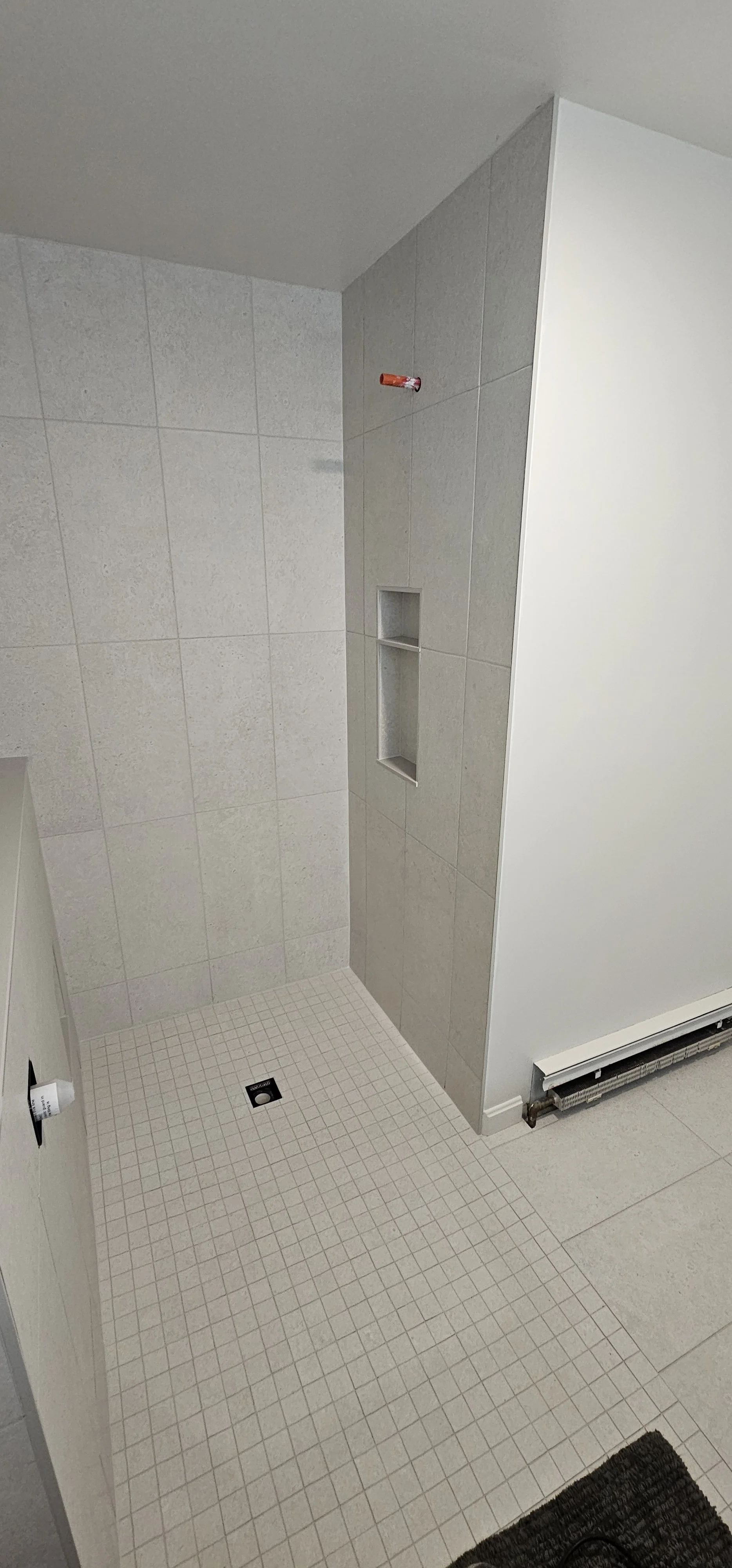 A bathroom shower area with beige wall tiles, a built-in niche, a drain on the floor, and a heating unit along the wall. The shower ceiling is not visible and there is no showerhead installed yet.