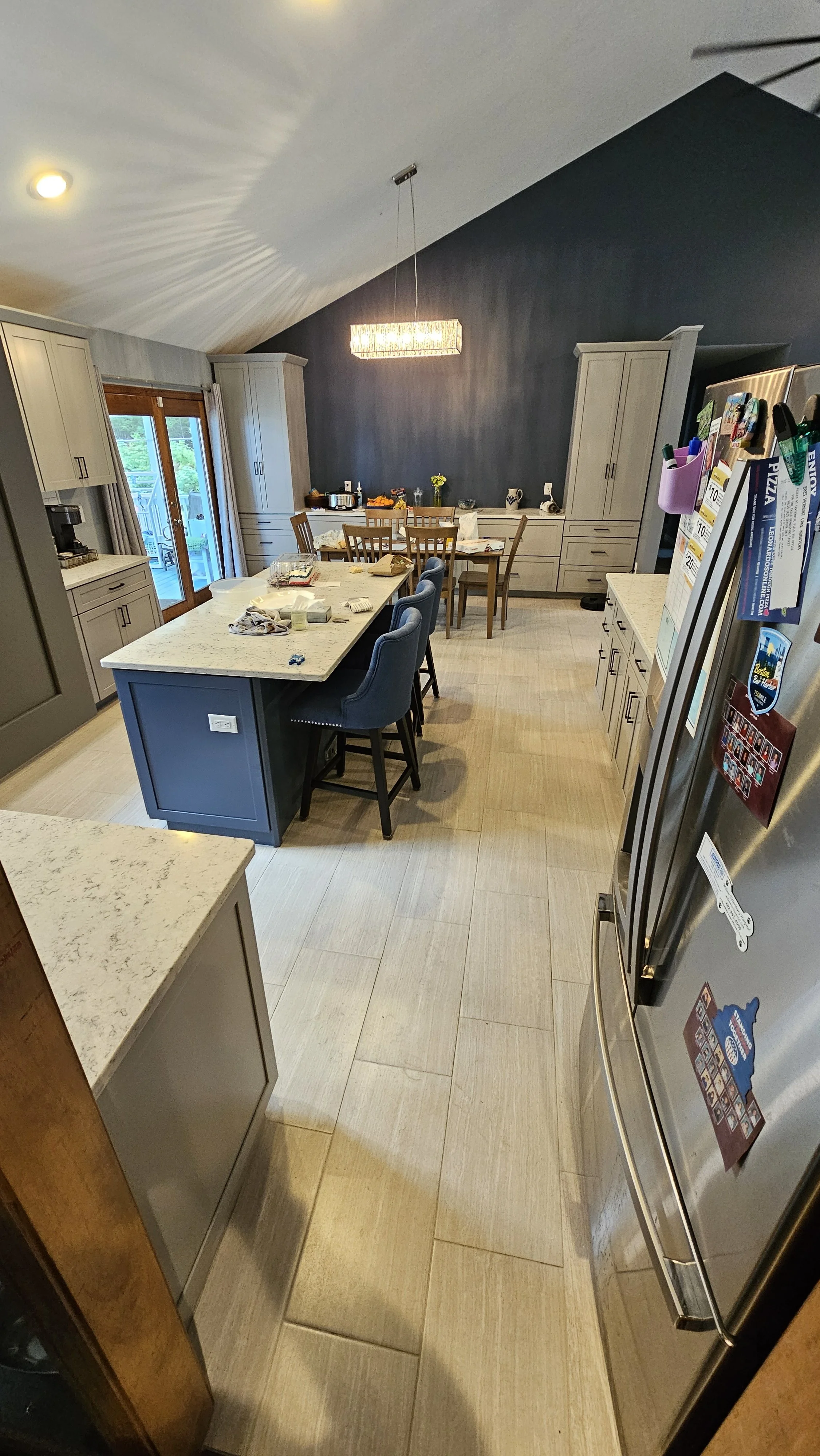 View of a modern kitchen with gray cabinets, a white island with blue chairs, a stainless steel refrigerator, and a dining area in the background featuring a dark accent wall, a rectangular light fixture, and a farm-style table with chairs. Sliding g