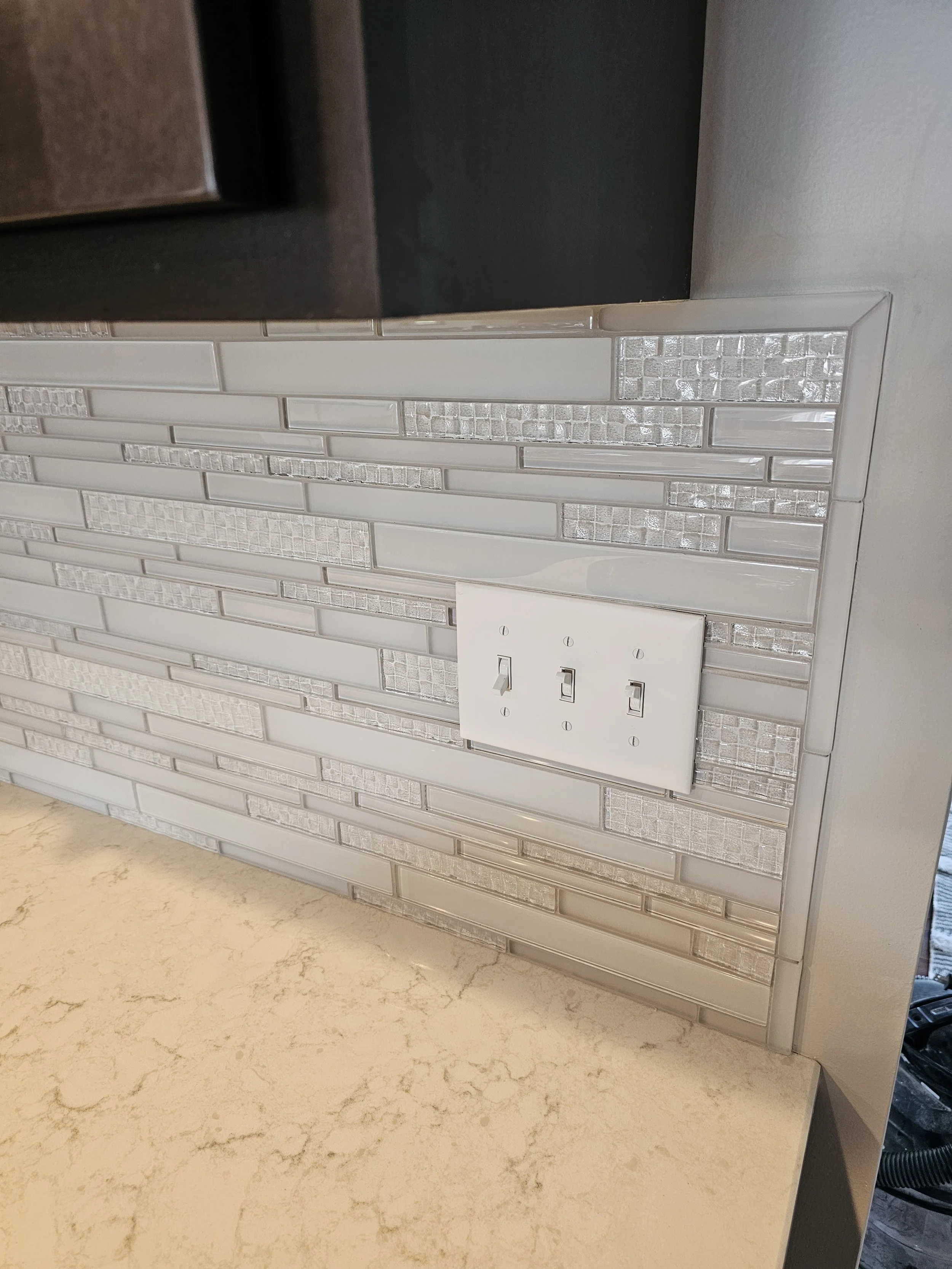 Close-up of a kitchen backsplash with white and textured glass tile, a white electrical outlet, and a beige marble countertop.