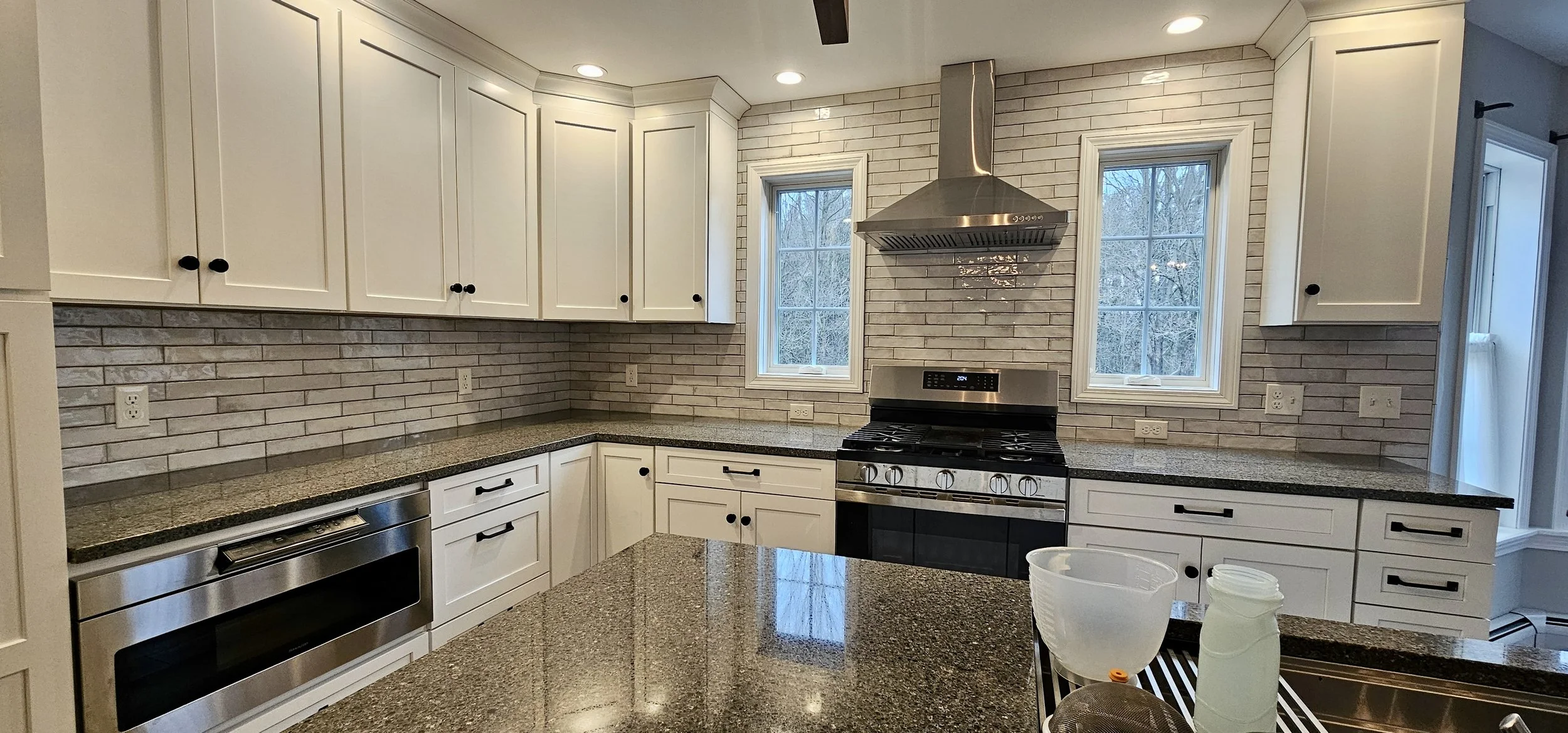Modern kitchen with white cabinets, granite countertops, and a brick backsplash. Features a stainless steel stove with an oven and a range hood, two windows, and various electrical outlets. Part of a kitchen island or counter is visible with some kitchen items.