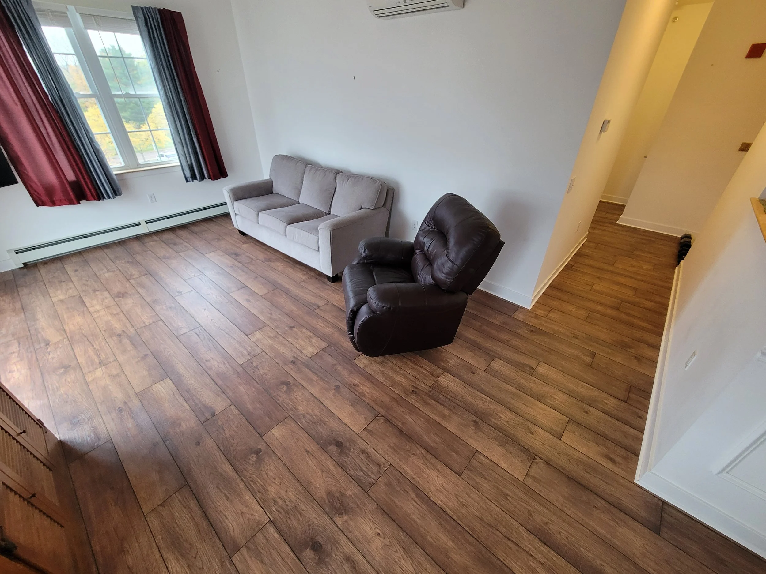 Living room with hardwood floors, a large window with red and navy curtains, a beige sofa, a black leather armchair, and a thermostat on the wall.