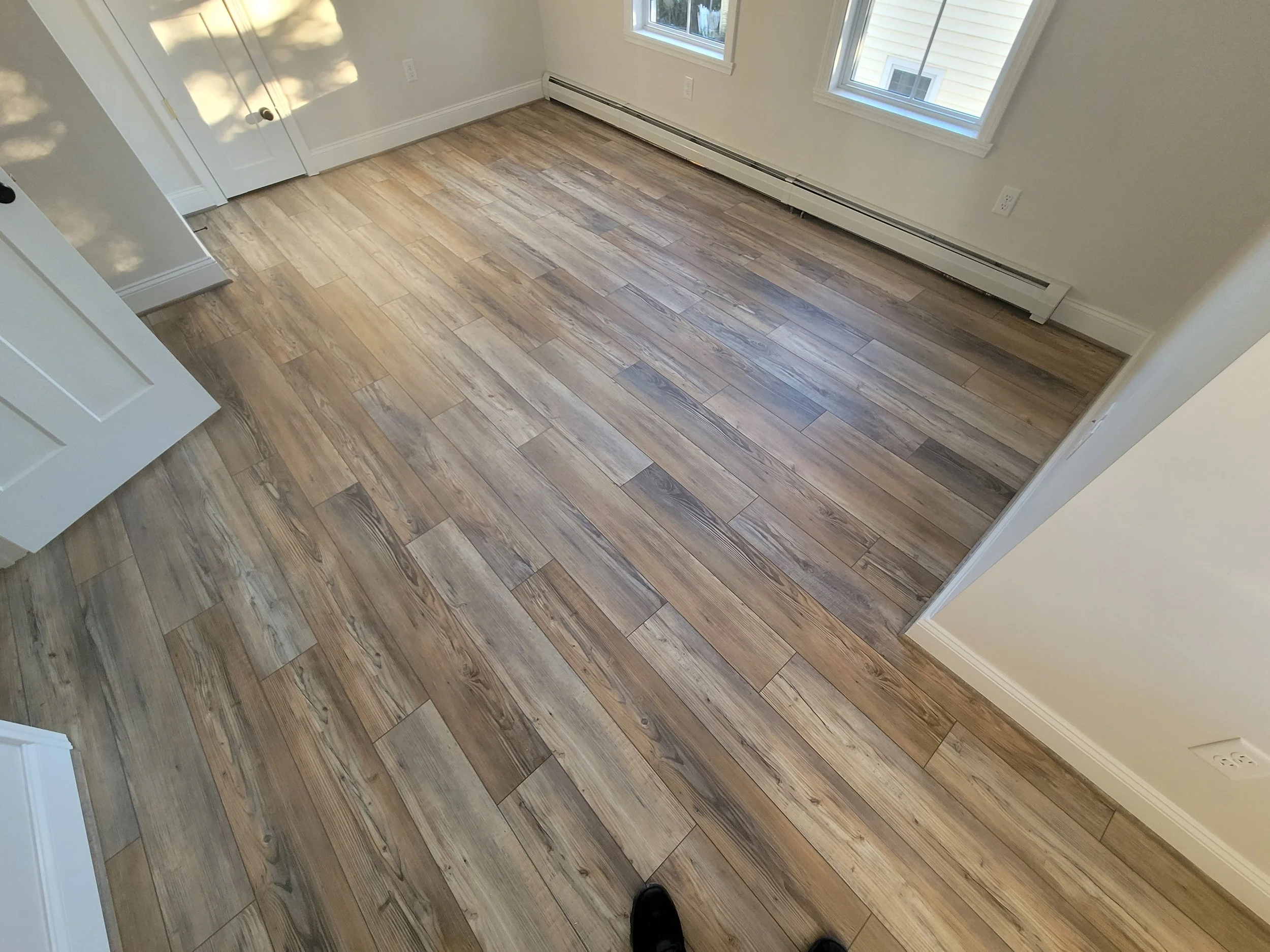A room with light-colored wood flooring, white walls, white window frames, and a white baseboard heater along the wall. Part of a white door with black doorknob is visible on the left, and sunlight is casting shadows through the window.