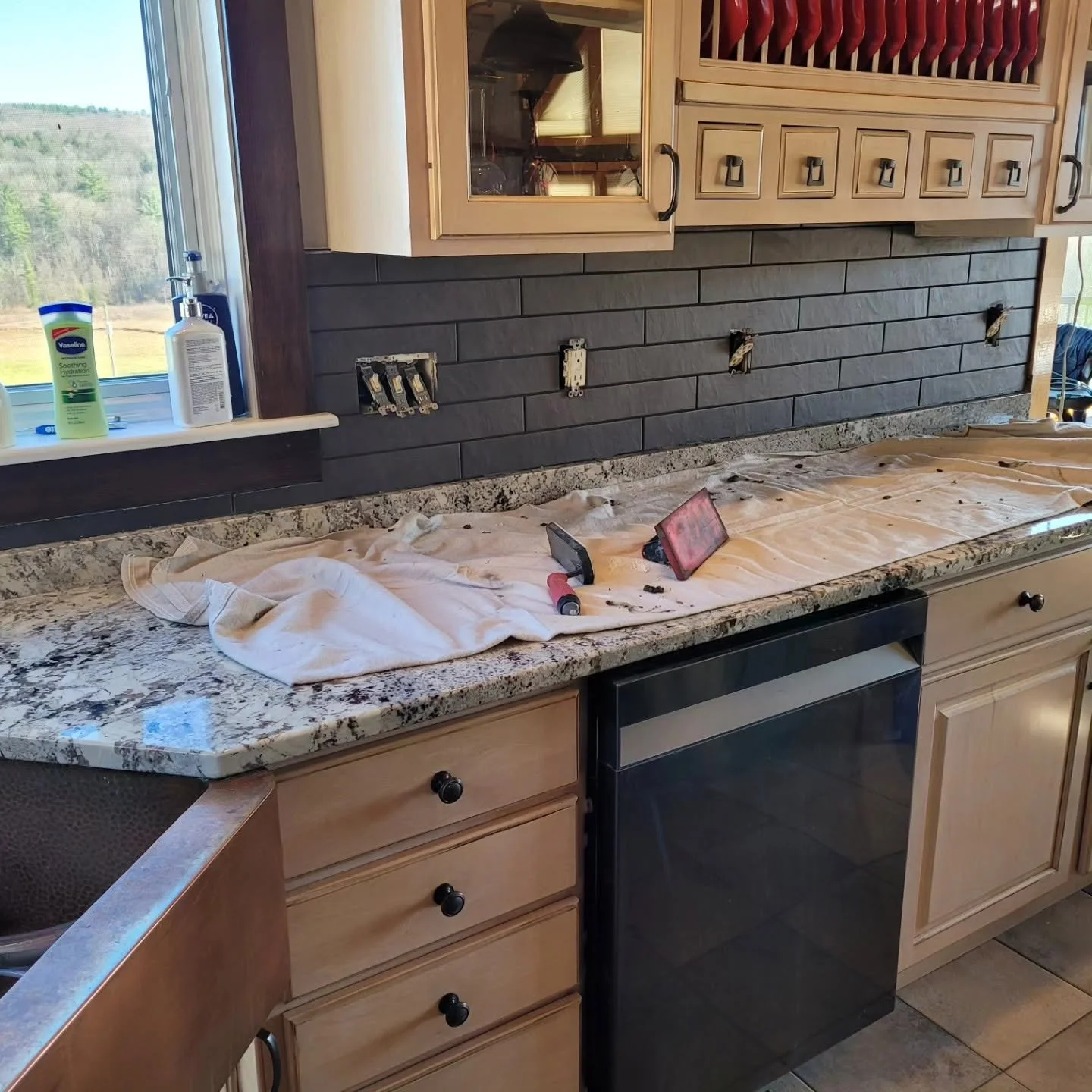 Kitchen counter with missing backsplash tiles, tools, and a cloth, next to a window showing an outdoor landscape.