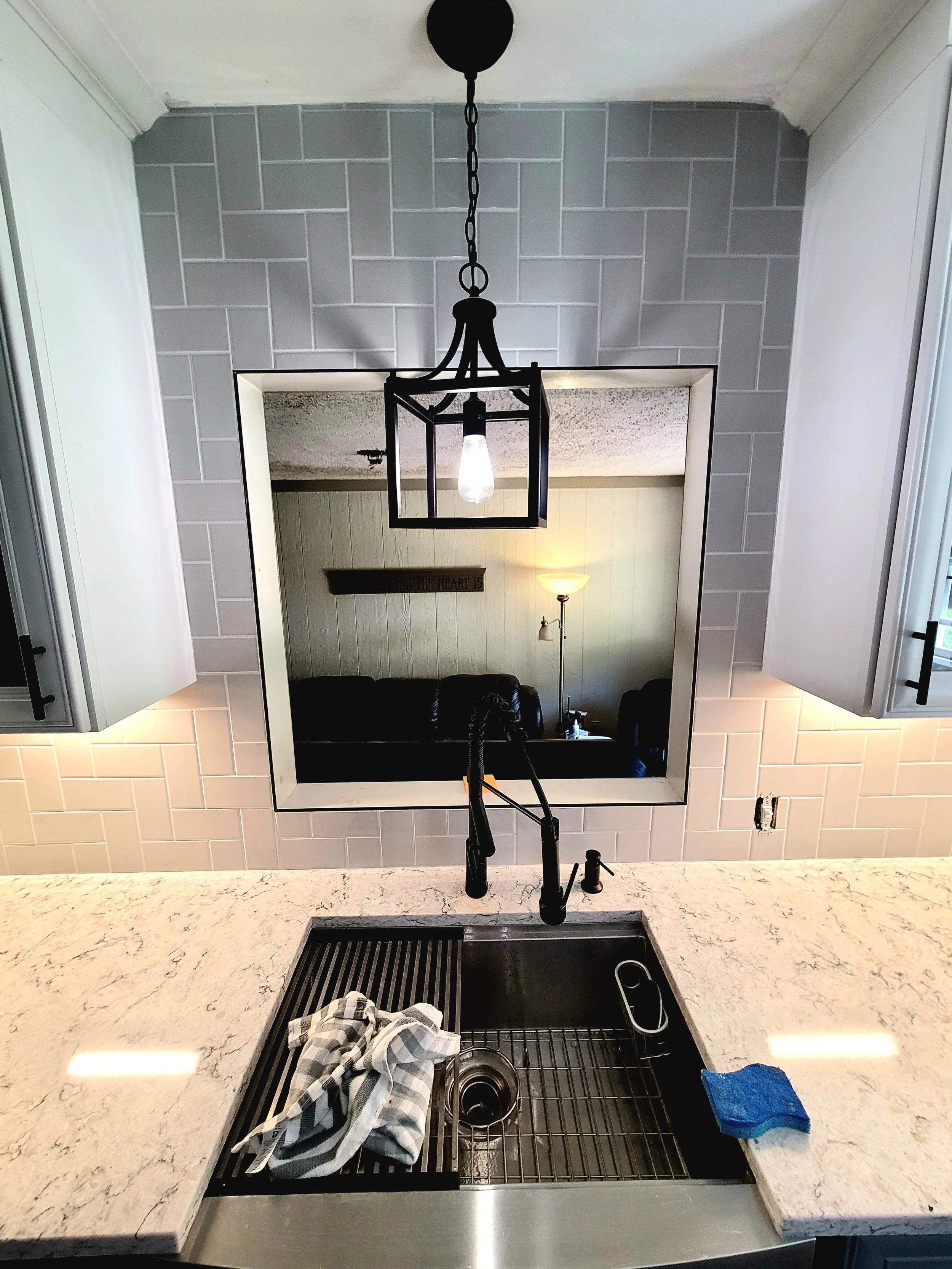 View from the kitchen countertop showing a sink with a dishcloth and scrubber, a black strainer in the sink, a faucet, and a mirror reflecting the living room with a wall sign and a standing lamp. The backsplash has white subway tiles, and there are 