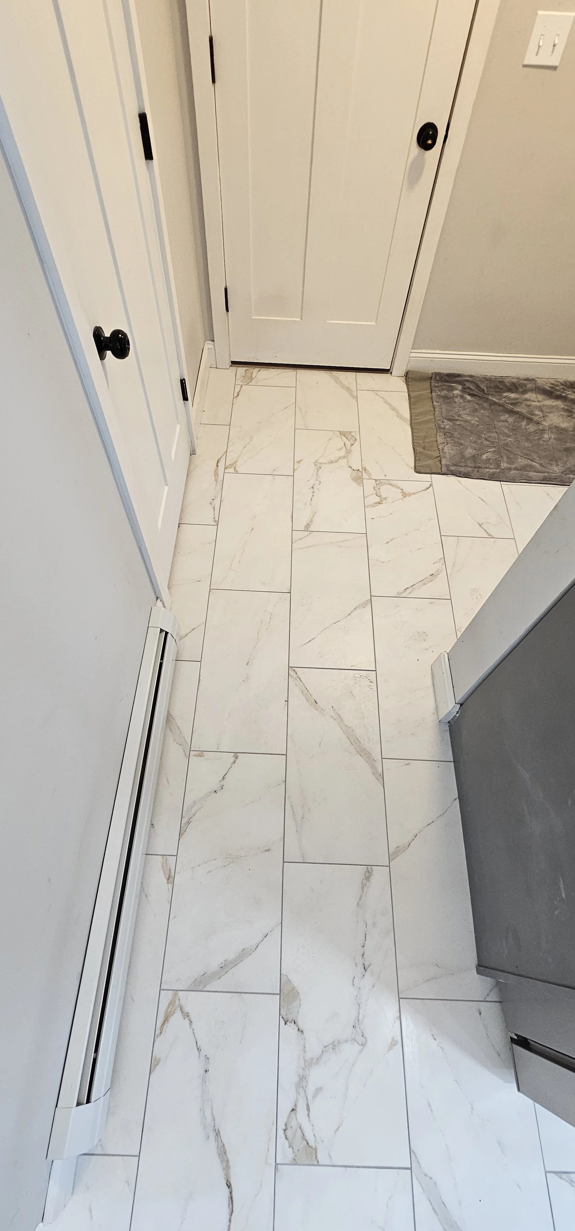 A hallway with white marble tile flooring, a white door, and a small gray rug near the door.