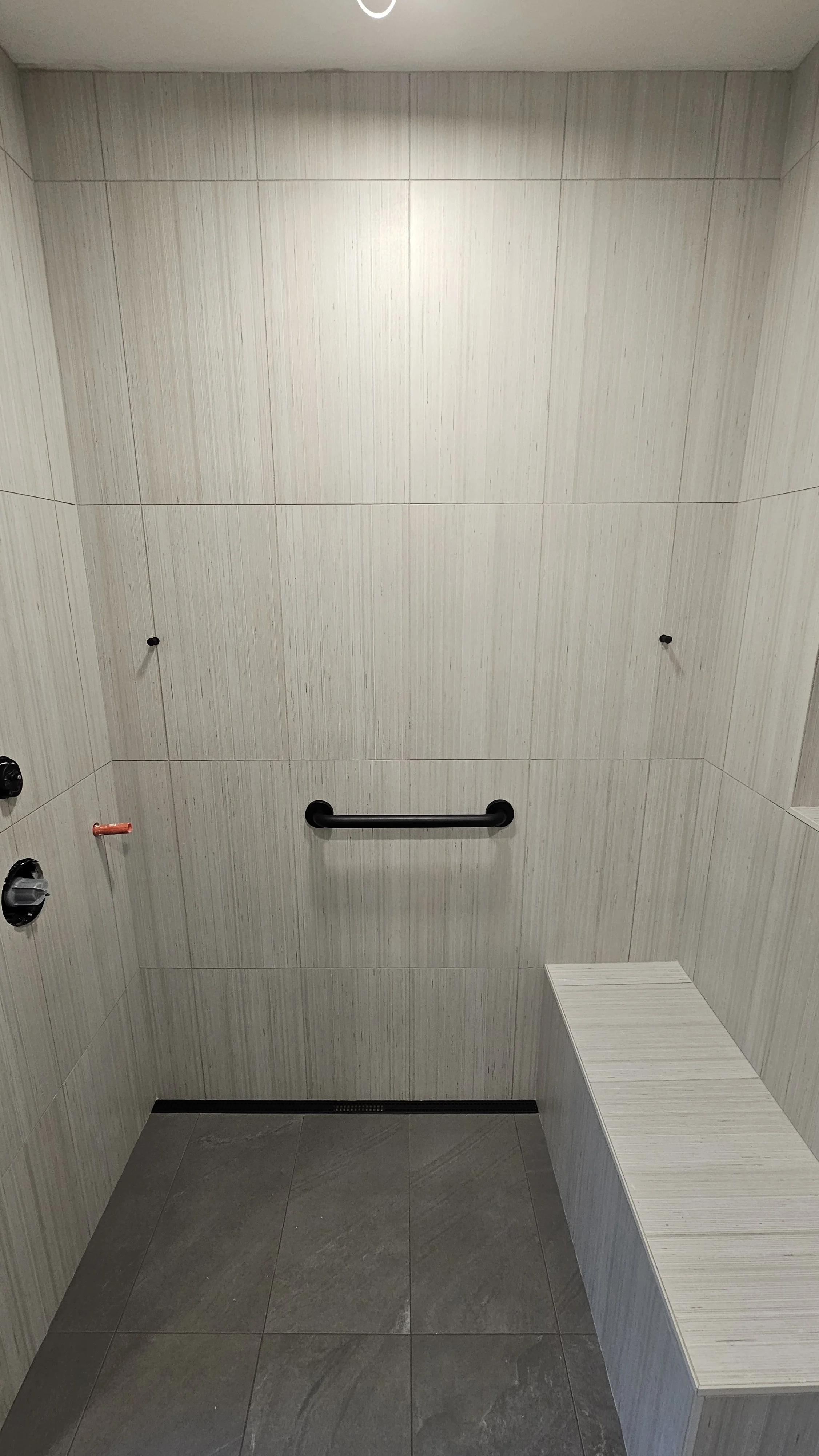 Interior of a shower stall with beige tiled walls, gray tiled floor, black grab bar, and a built-in bench on the right side.