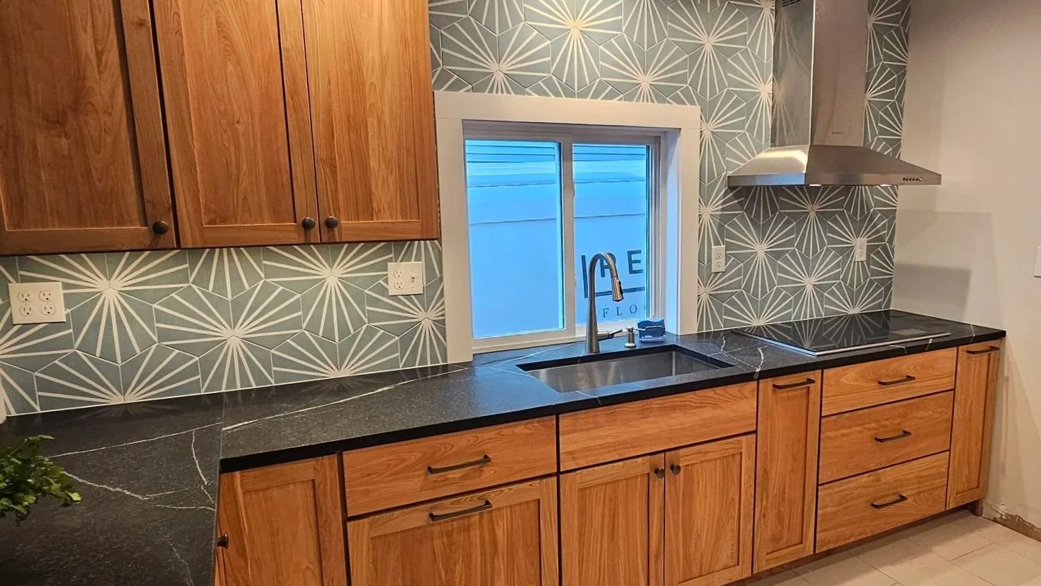 Kitchen with wooden cabinets, a black countertop with white veins, a window above the sink, a stainless steel range hood, and a geometric patterned backsplash.