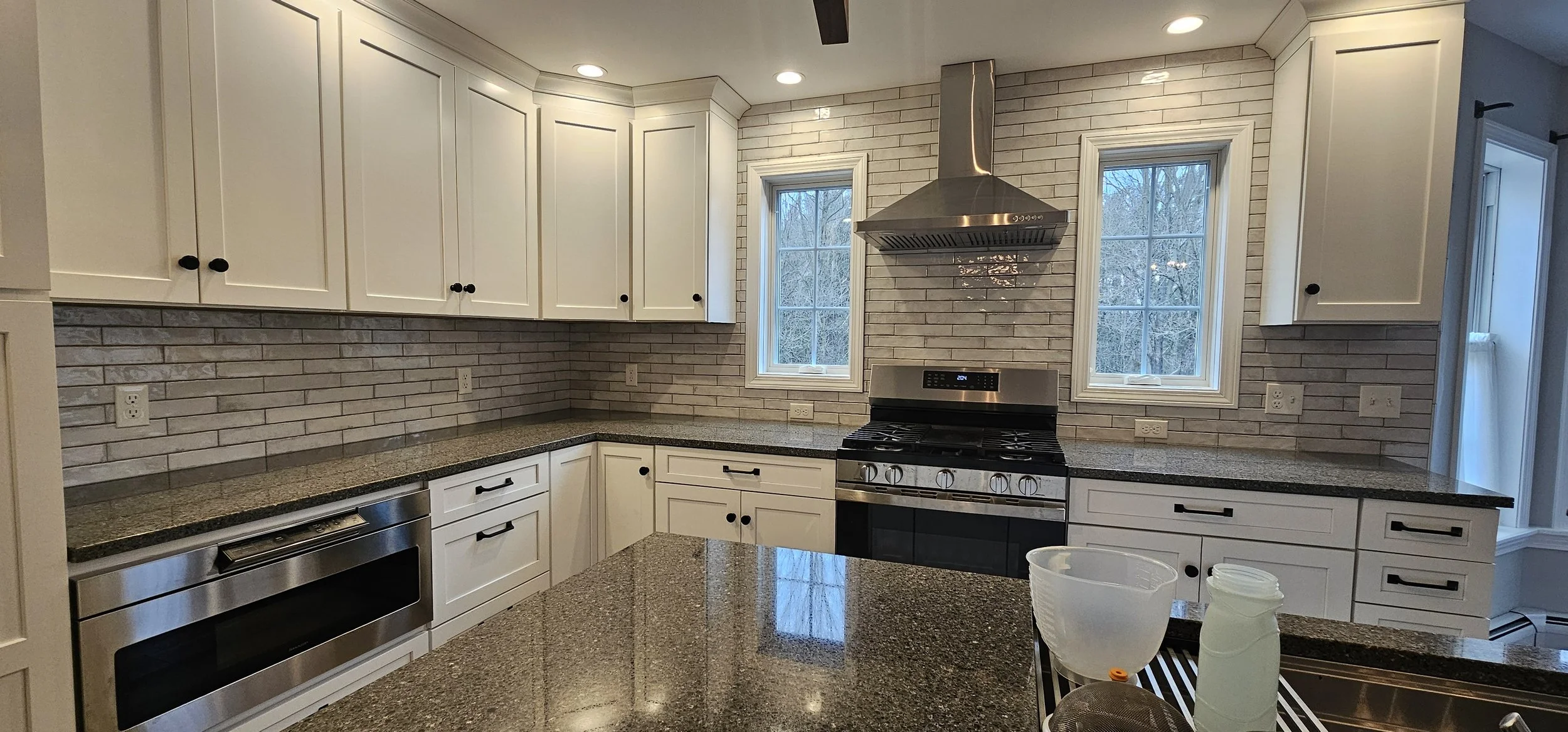 Modern kitchen with white cabinets, granite countertops, brick backsplash, stainless steel appliances, and two large windows showing a view of trees outside.
