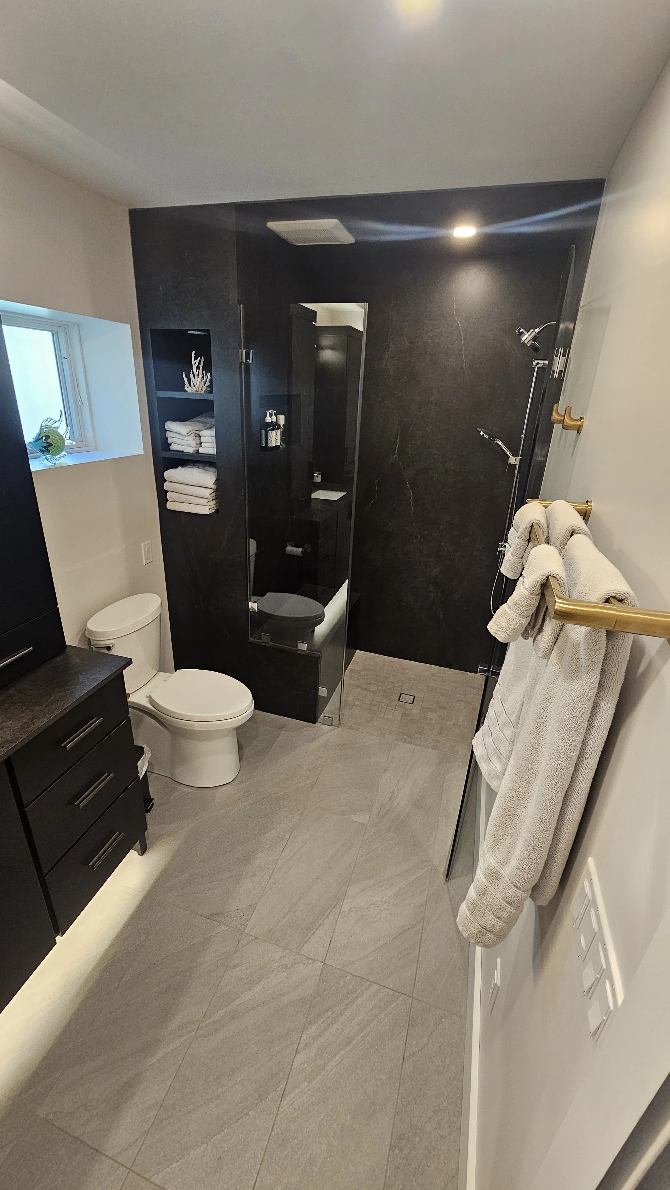 Modern bathroom with gray tiled flooring, a black vanity with drawers, a white toilet, a walk-in shower with glass enclosure and black walls, a small window with decorative glass, and beige towels hanging on a gold towel rack.
