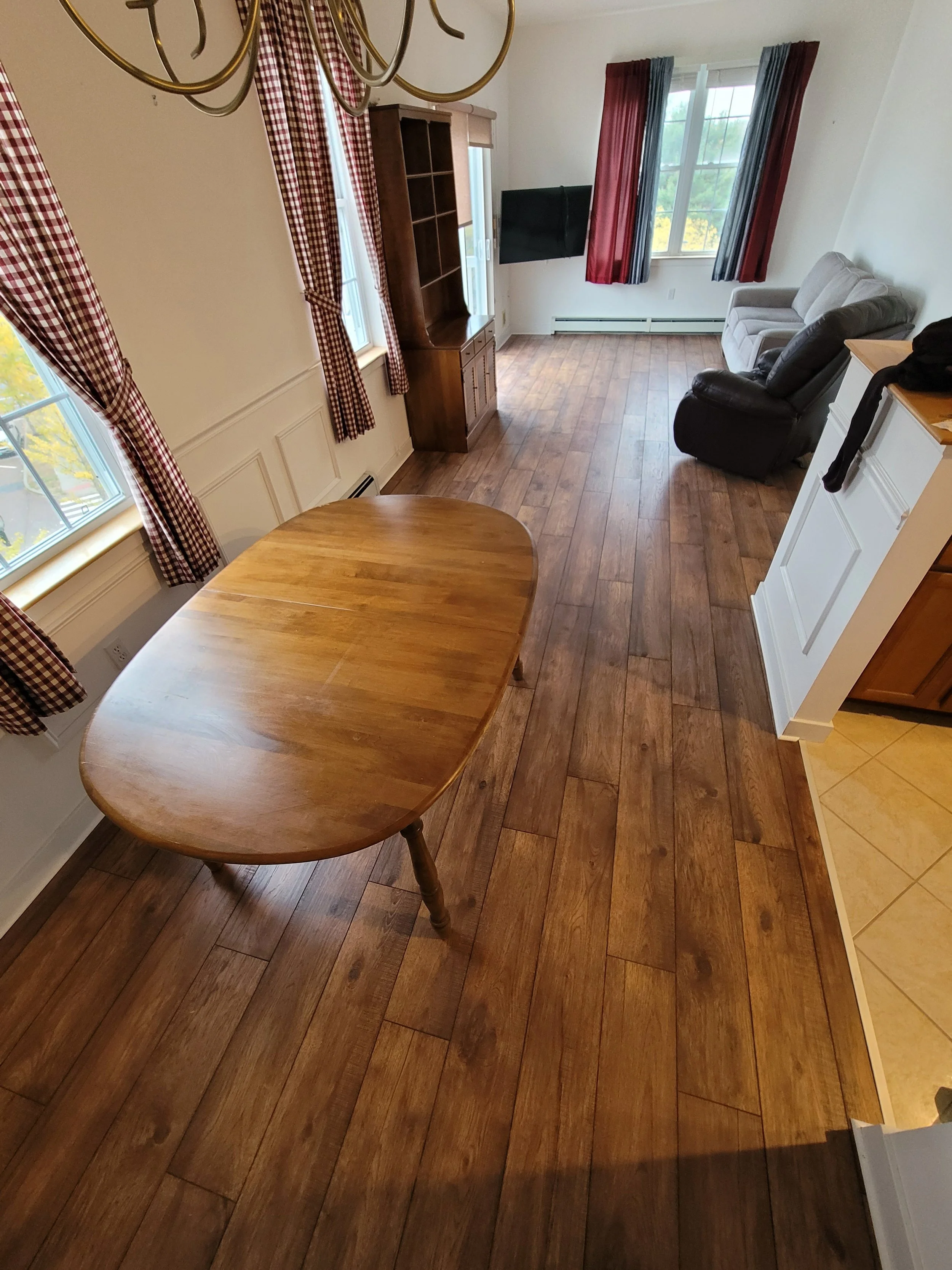 The image depicts a cozy living room area with wooden flooring, a wooden dining table in the foreground, and a cozy seating area with two recliners and a sofa near the window. The room has large windows with red plaid curtains and blue curtains, allo