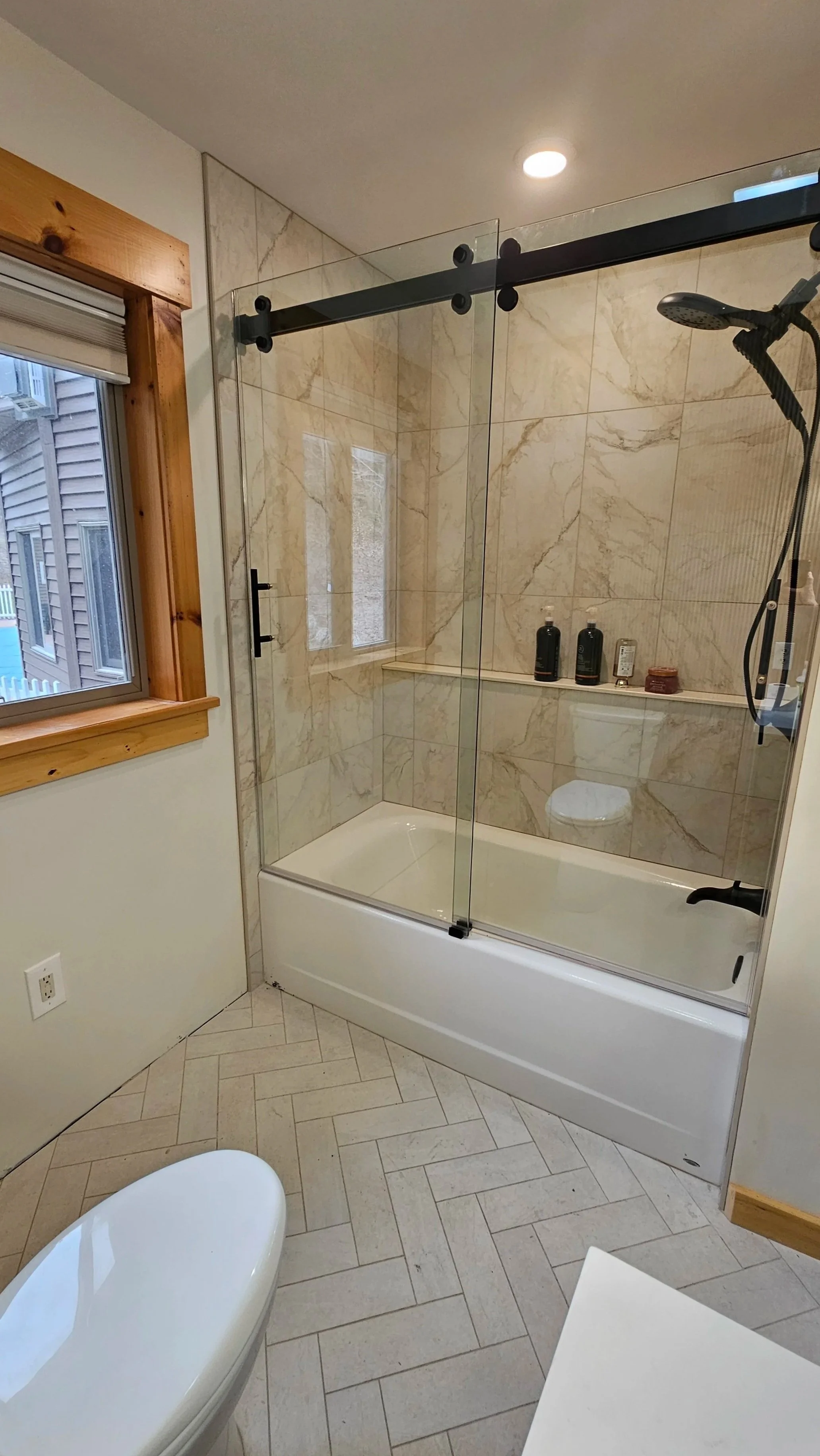 Bathroom with walk-in shower with glass doors, a window with wooden trim, a white toilet, and beige tiled walls and floor.