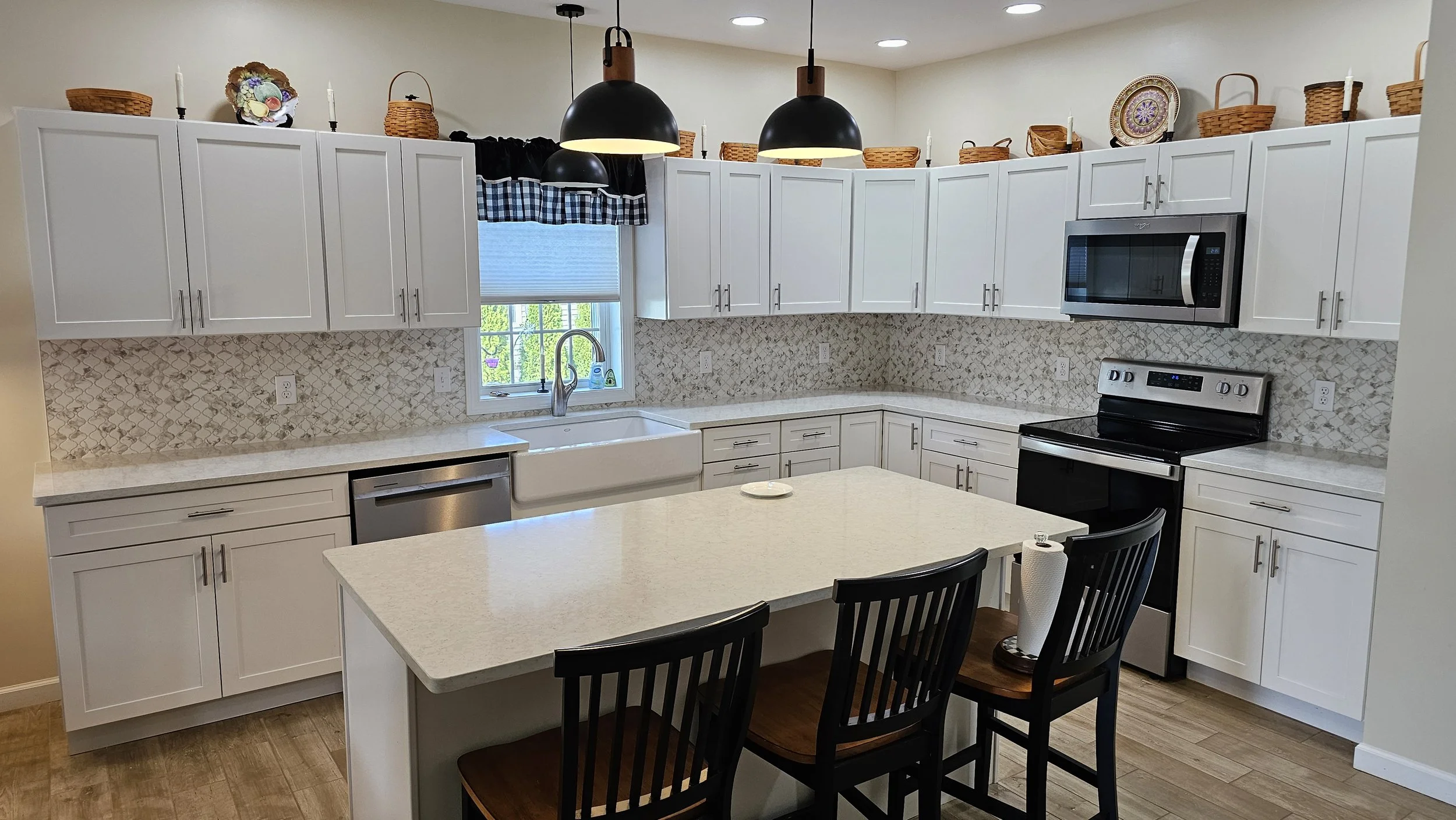 A modern kitchen with white cabinets, a kitchen island, a window above the sink, black pendant lights, a microwave, a stove, and decorative baskets on top of the cabinets.