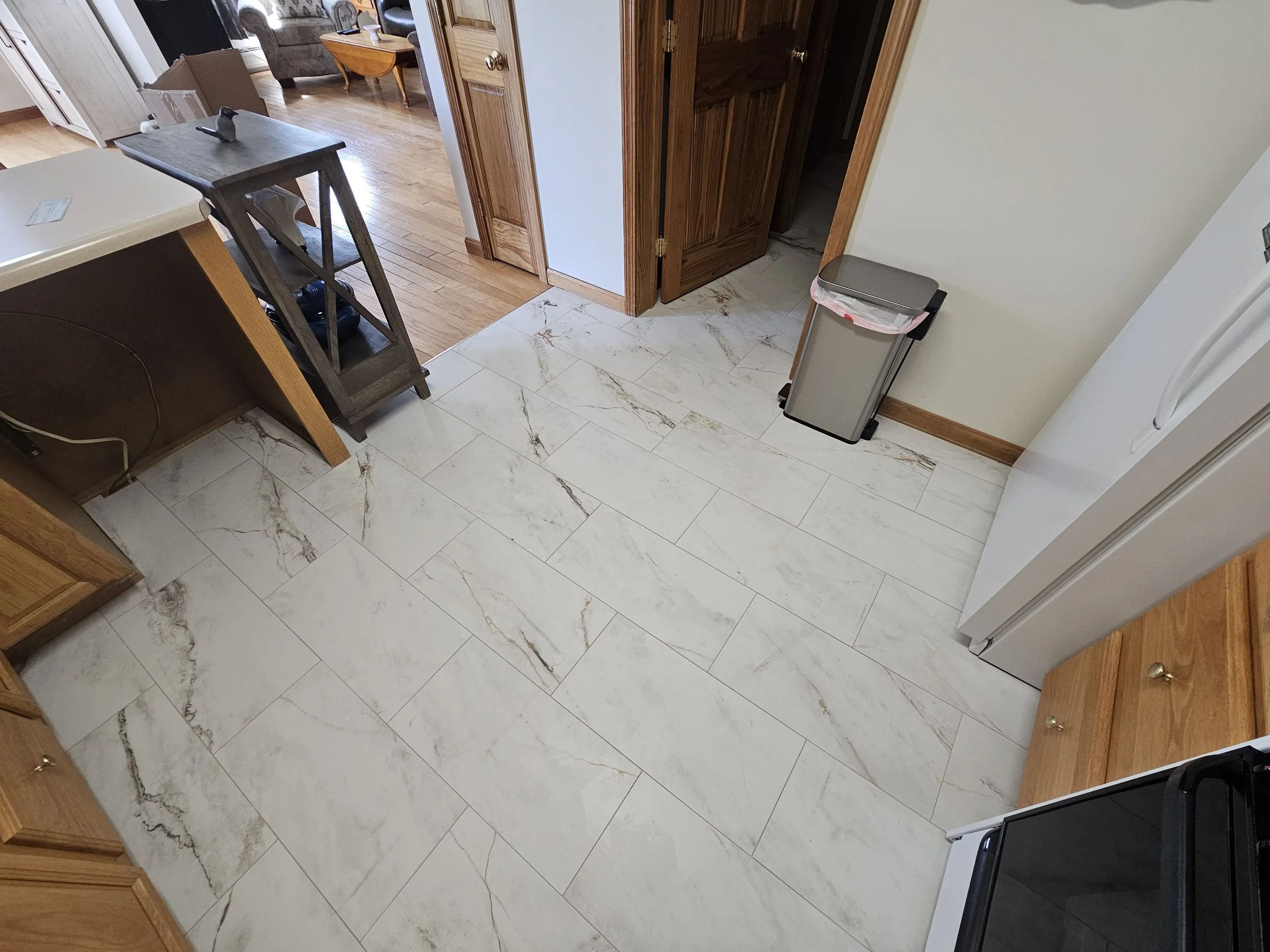 Interior view of a kitchen area with marble tile flooring, wooden cabinets, and a trash bin. The kitchen opens into a living space with hardwood flooring, a sofa, and a wooden coffee table.