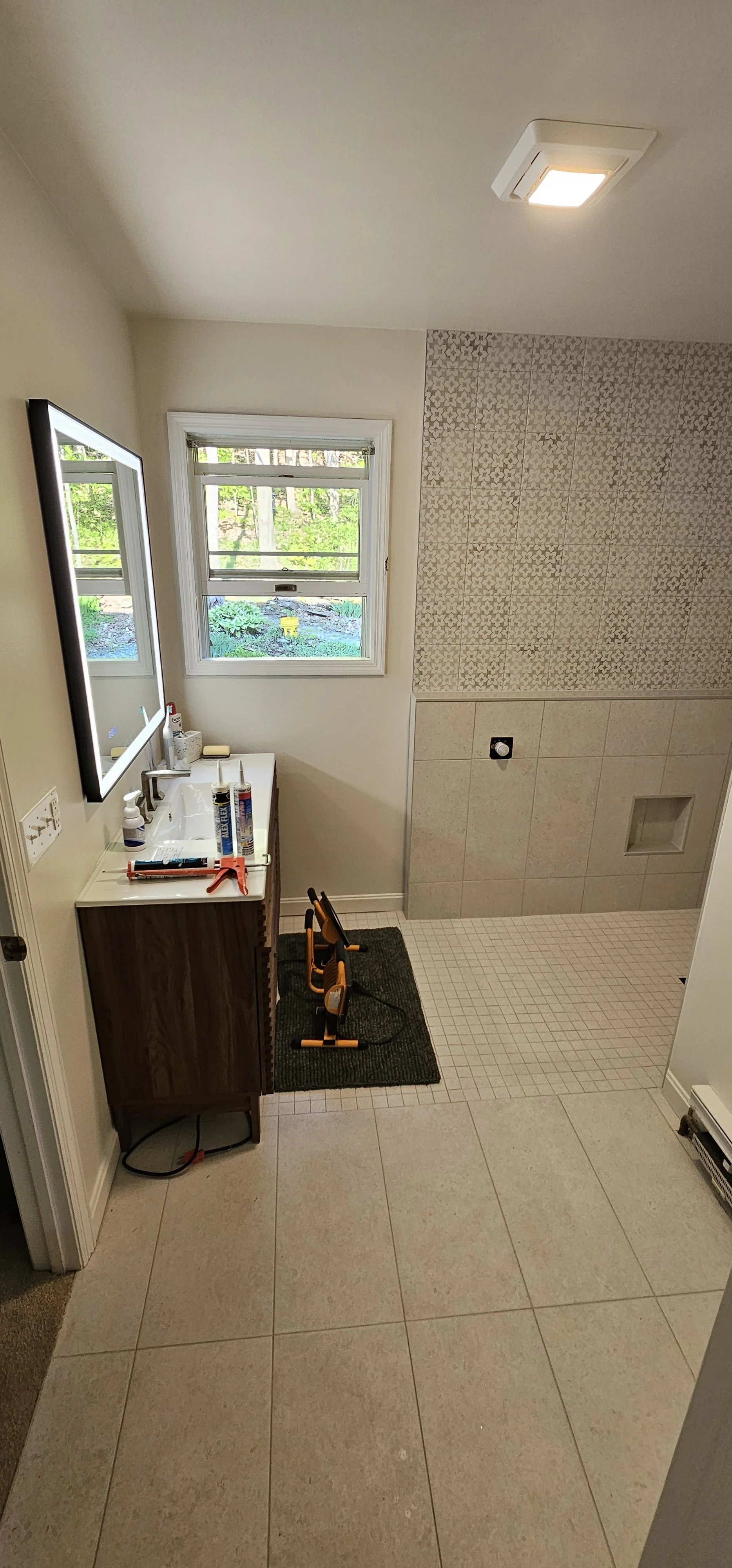 Bathroom with white tile floors, a vanity with a mirror, window with green view, shower area with beige and patterned tiles, and two small exercise bikes on a mat.
